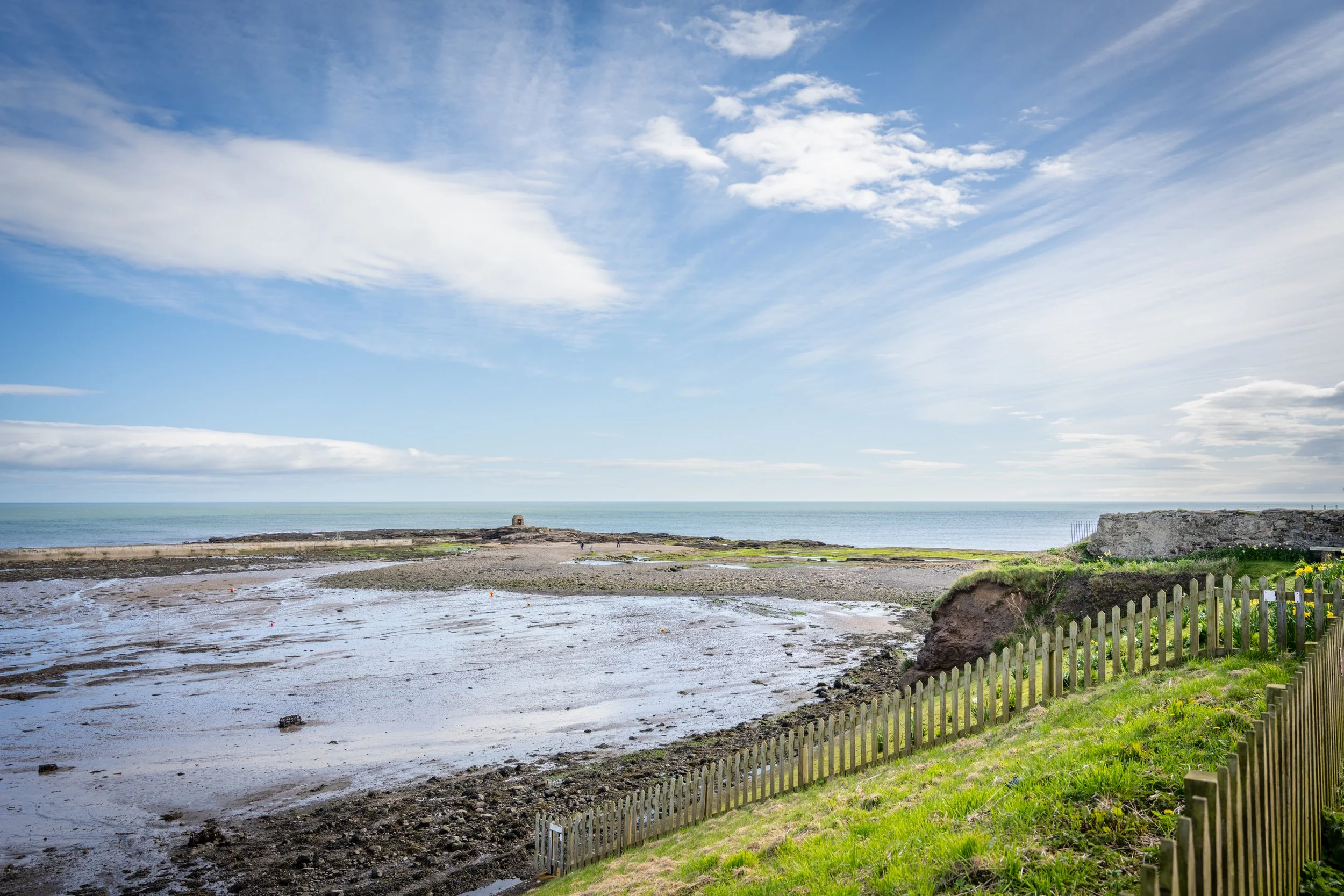 A scenic coastal view with a partly cloudy blue sky, calm ocean, rocky shoreline, and a small white lighthouse in the distance. Green grass and a wooden fence are in the foreground.