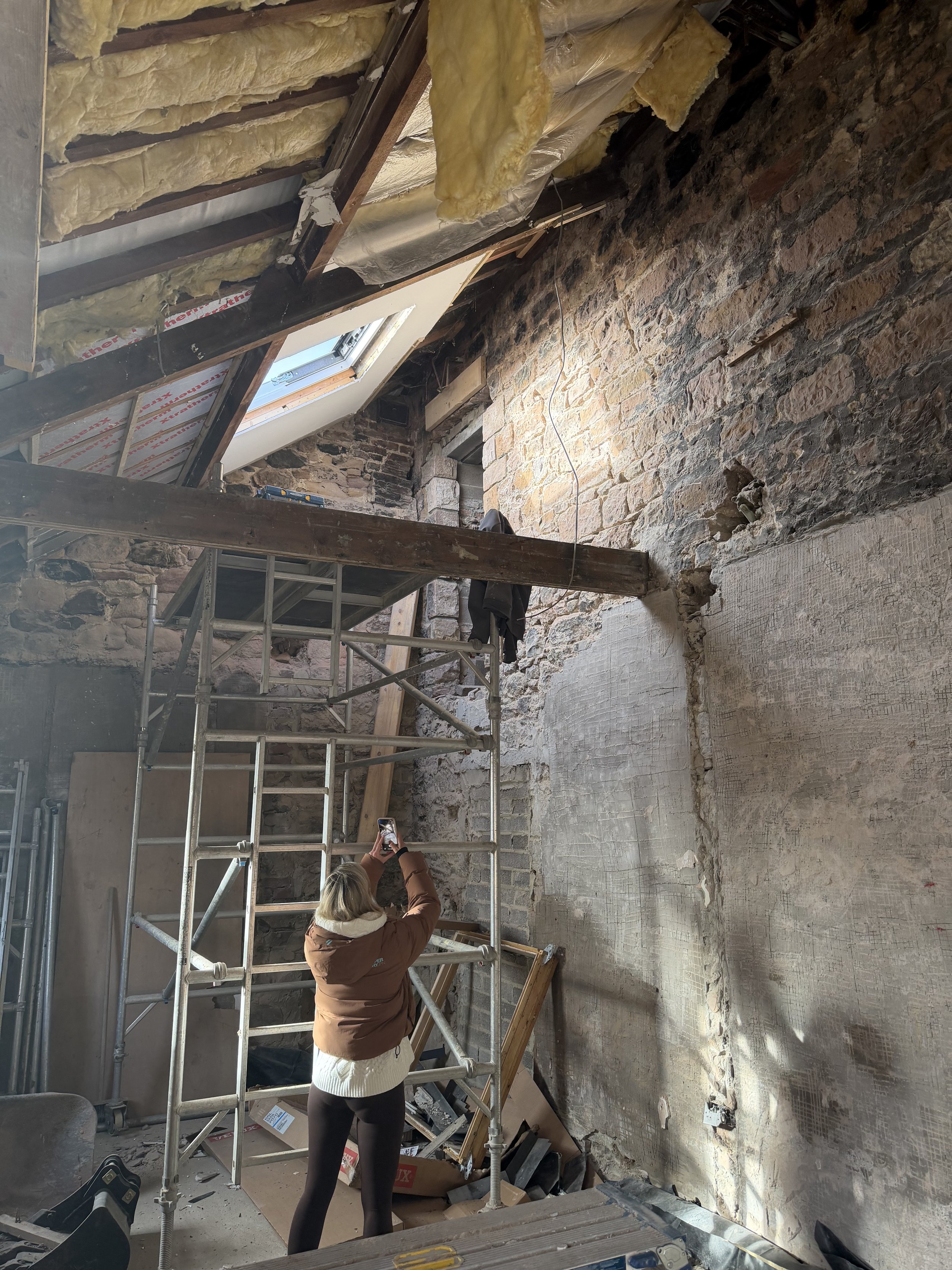 A woman taking a photo with her phone inside a room under renovation. The room has exposed brick walls, a sloped ceiling with insulation and insulation boards, and scaffolding for construction work.