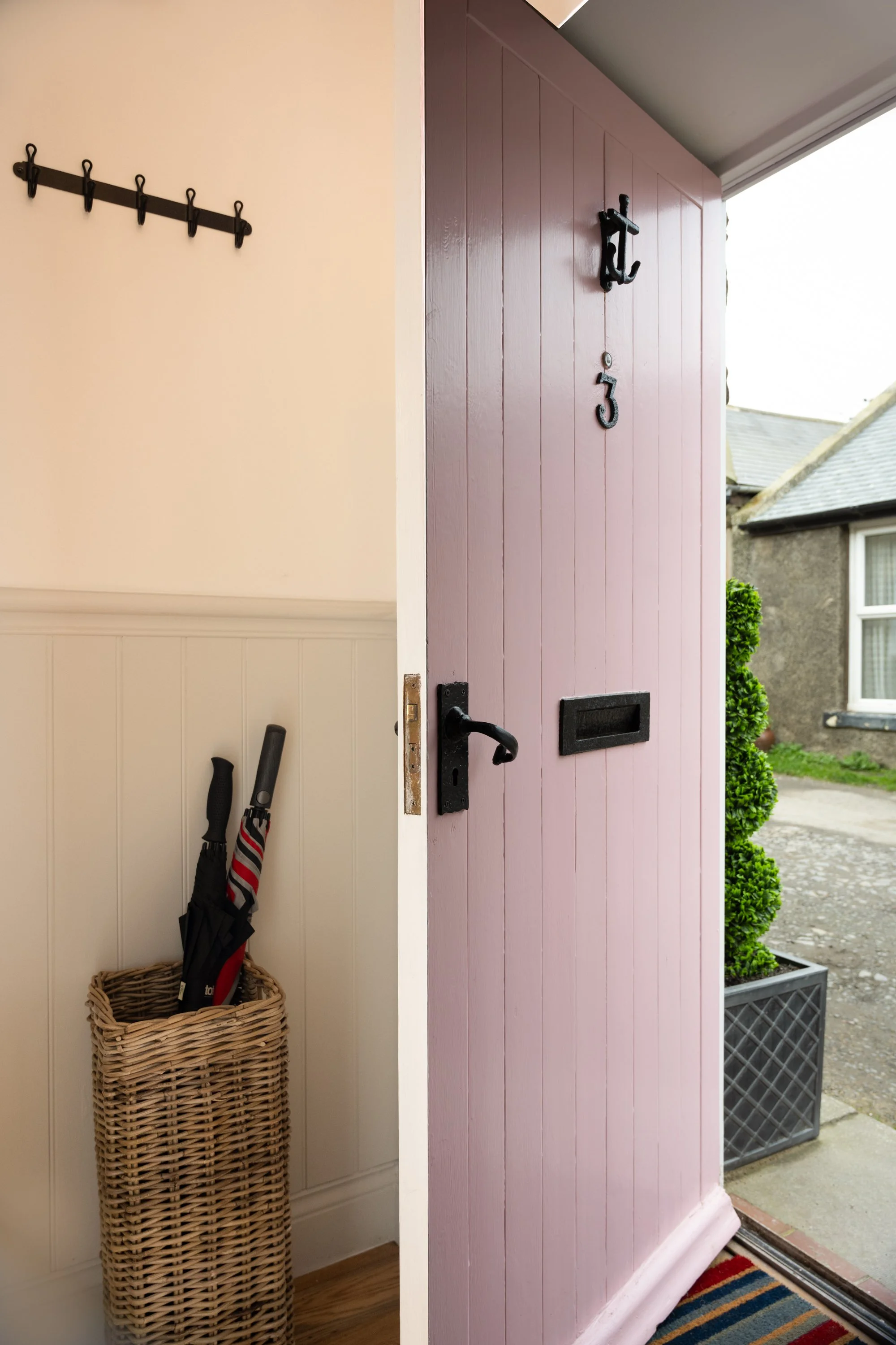 Entrance door with house number 133, partially open, showing a glimpse of the outside with a small garden and neighboring house. Inside, a basket with umbrellas and a wall rack with hanging umbrellas can be seen.