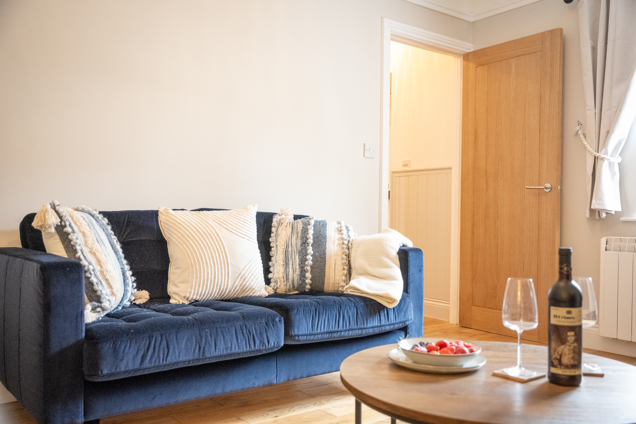 A living room with a navy blue sofa decorated with beige and black pillows, a wooden coffee table with a bowl of red tomatoes, bottles of wine, and glasses, beige walls, a wooden door, and a window with white curtains.