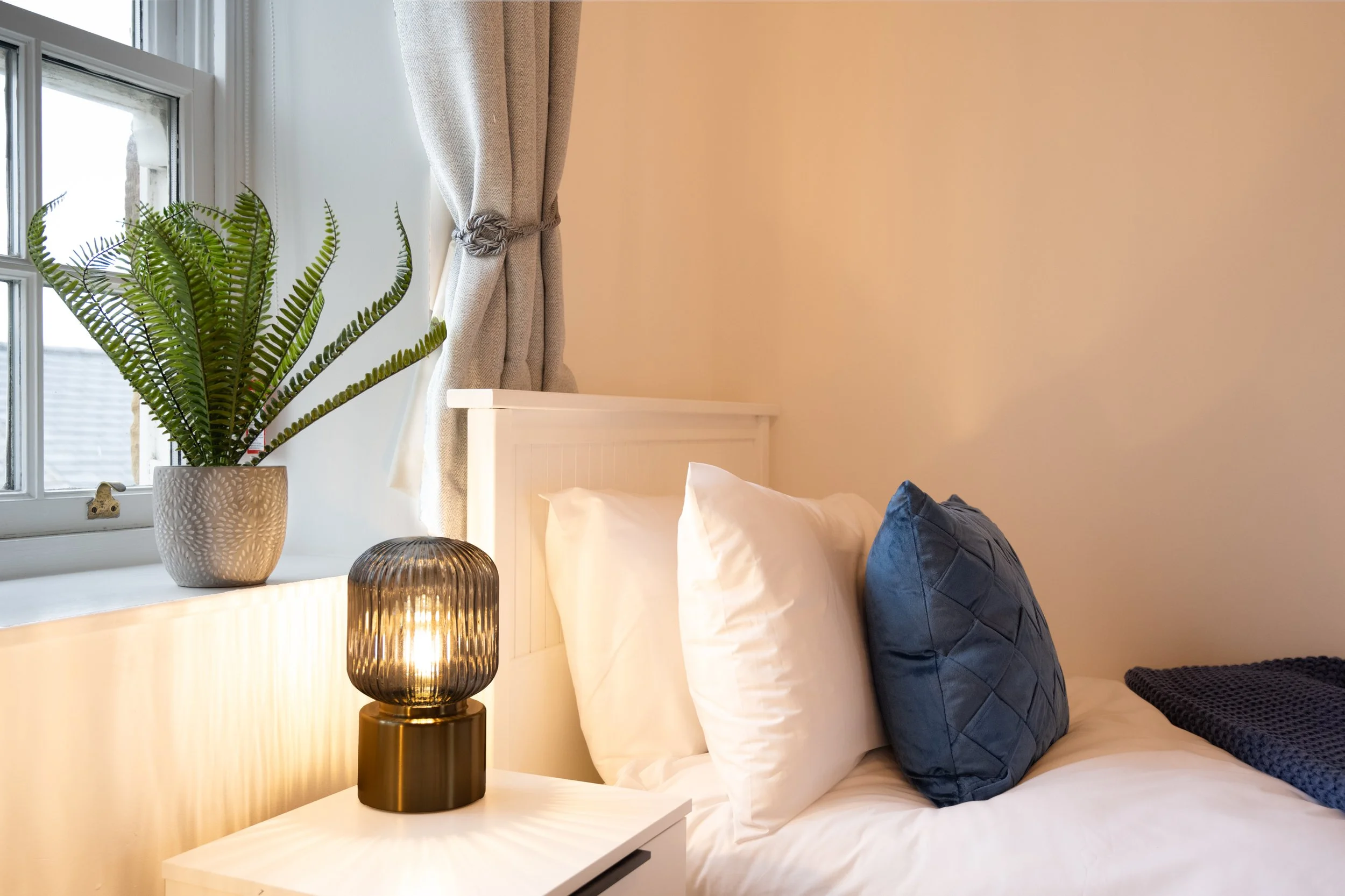 A bedroom corner featuring a white-painted wooden headboard, a large green fern in a decorative pot on the windowsill, a dark ribbed table lamp, and a window with curtains tied back, letting in natural light.