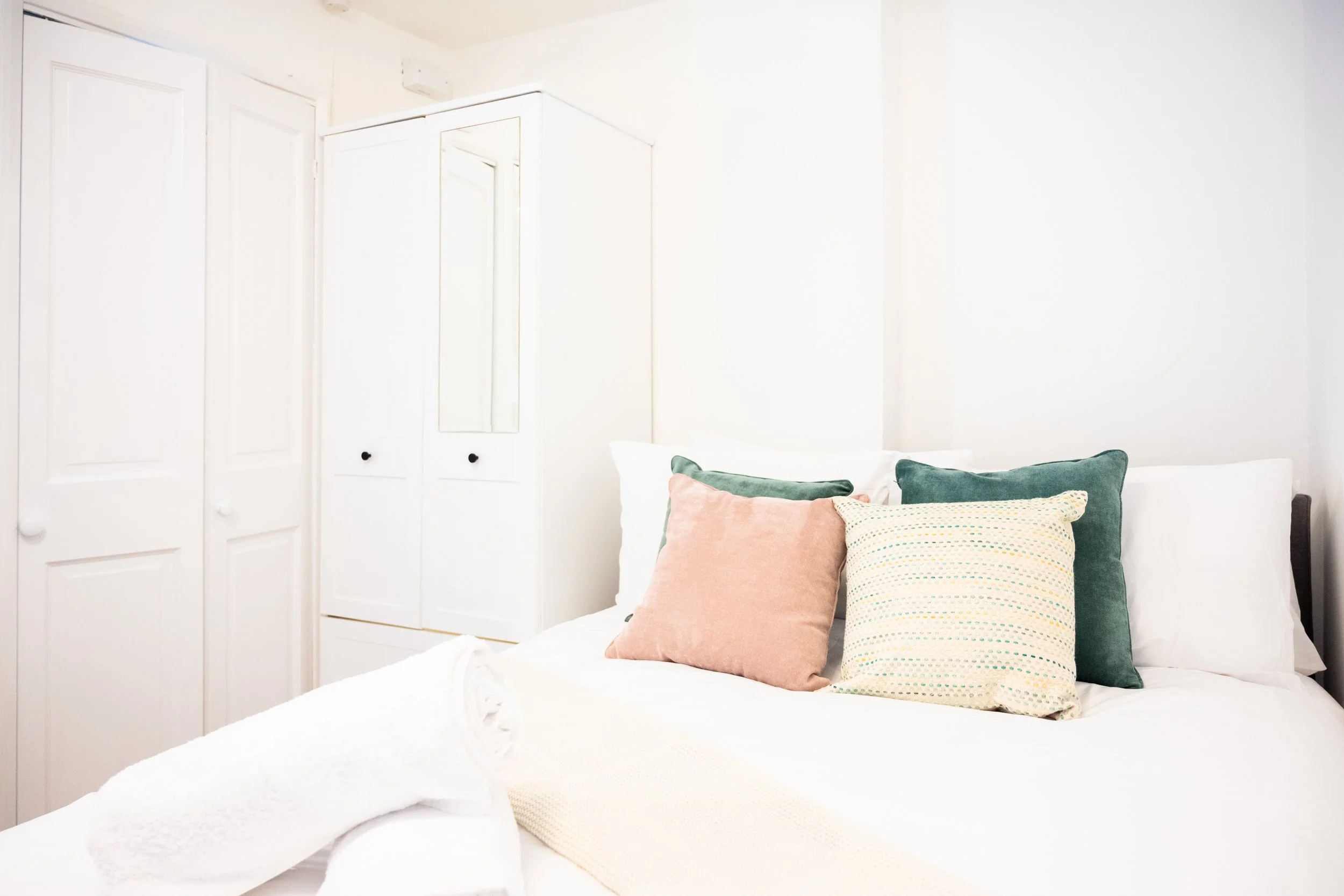 A neatly made bed with a white sheet and several pillows in pastel colors, including pink, green, and cream, in a cozy bedroom with white walls and a wardrobe with a mirror.