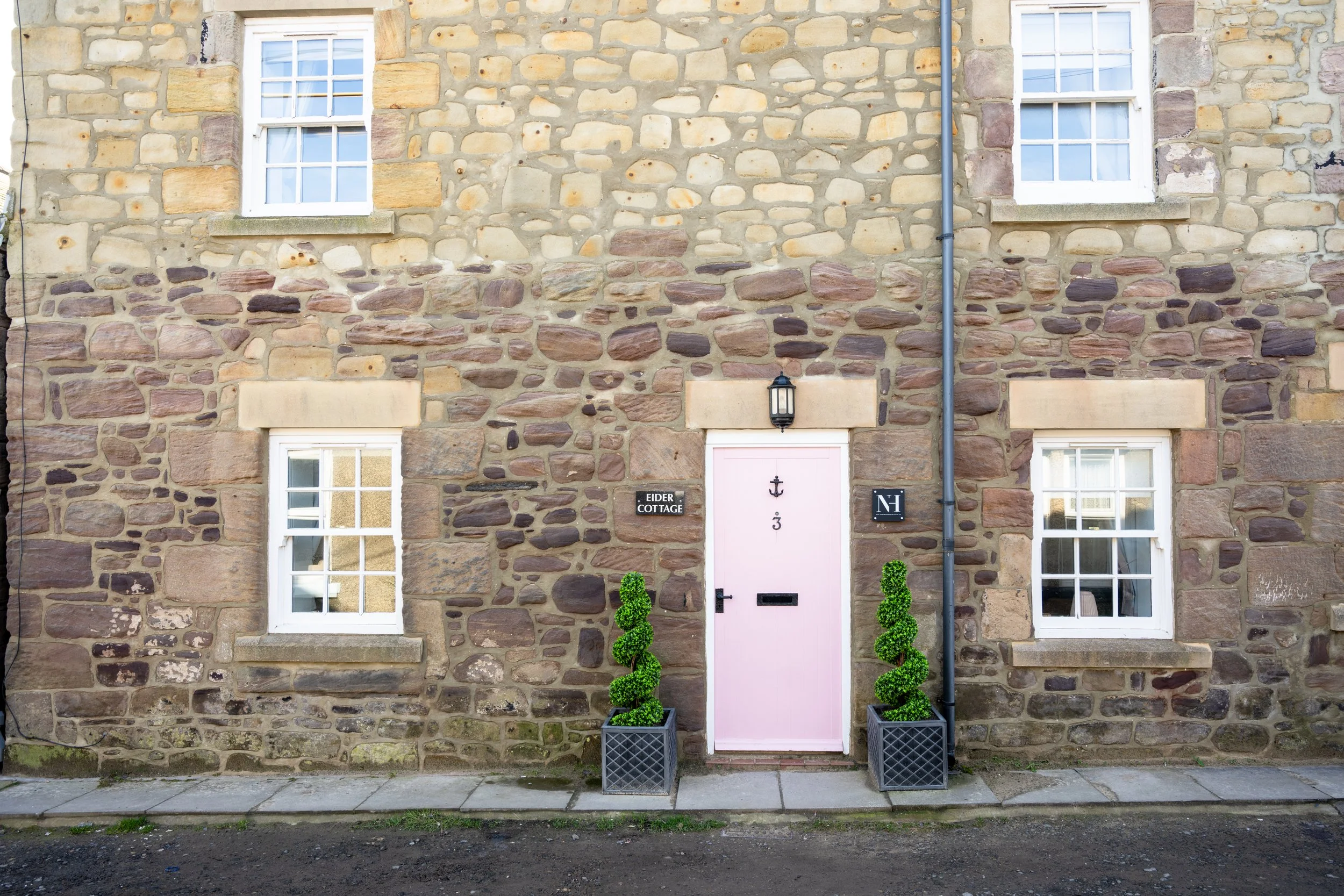 Photo of a stone cottage facade with two white-framed windows, a pink door with an anchor decoration, and two small green topiary plants in black rectangular planters, with a black vintage-style wall lamp and signs reading 'EIDER COTTAGE' and 'N' nea