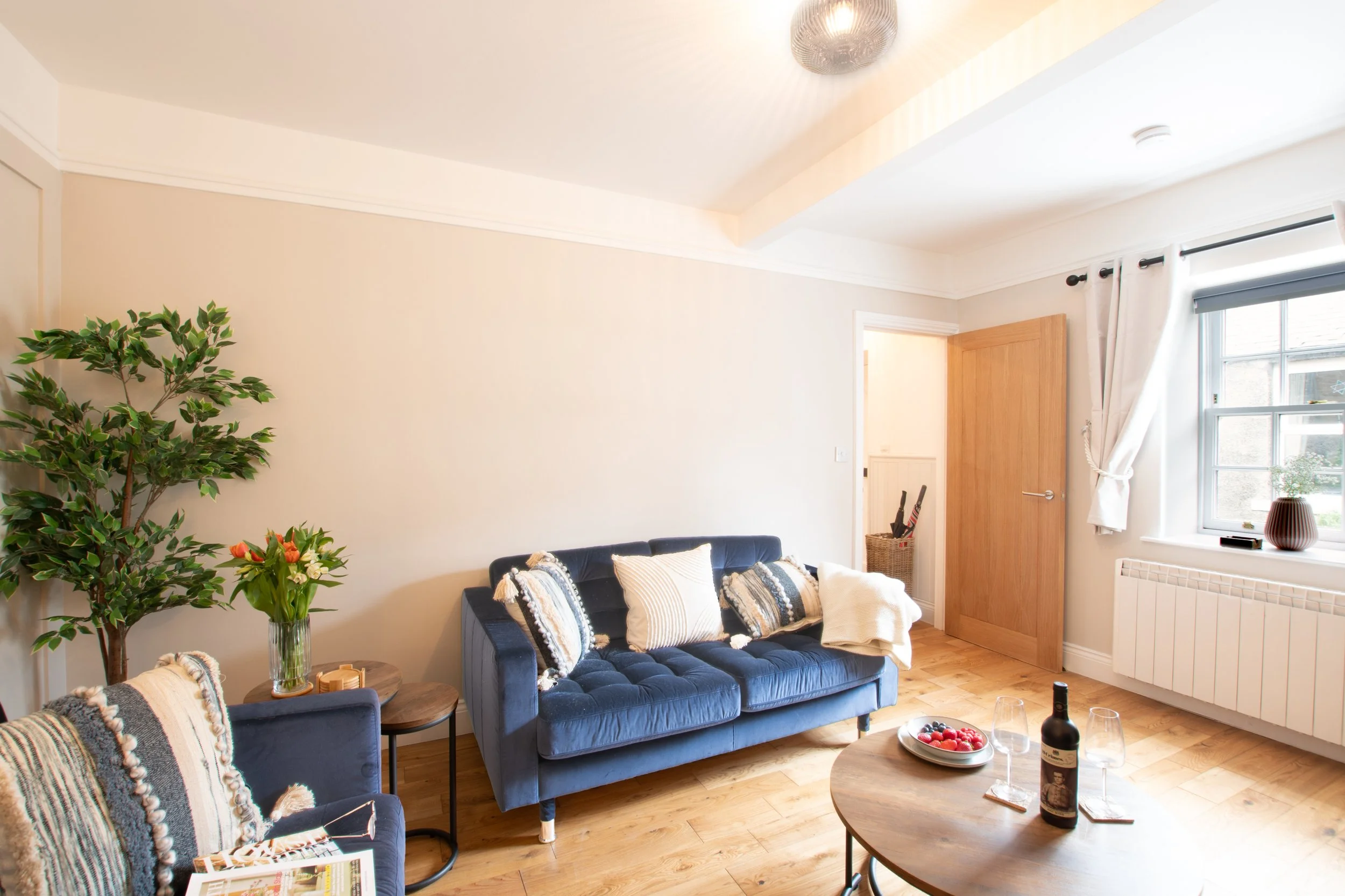 Living room with beige walls, wooden flooring, a navy blue couch, decorative pillows, a side table with a book and vase of flowers, a coffee table with a bottle of wine, glasses, and a bowl of strawberries, a large window with white curtains, a potte