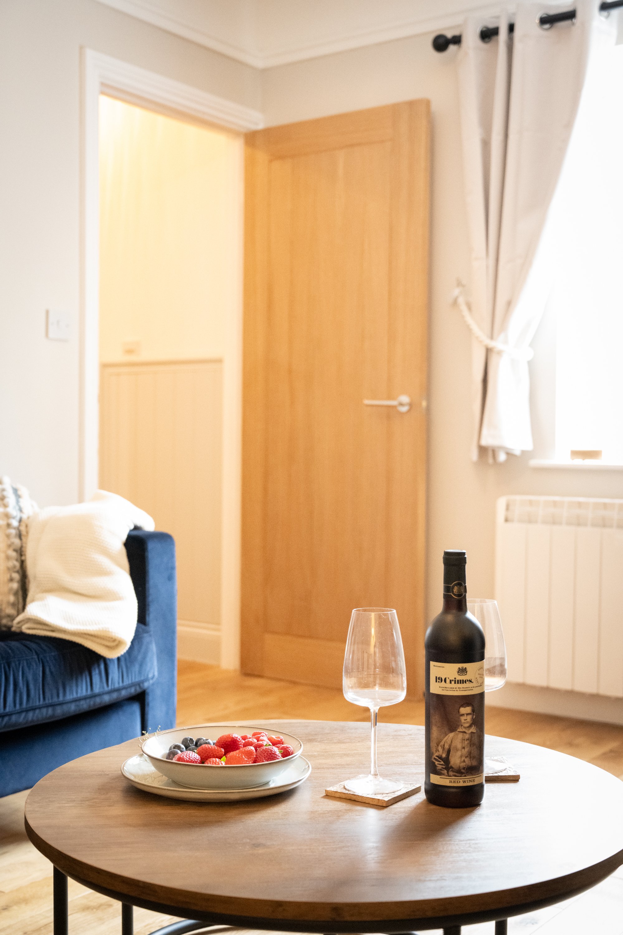 A round wooden table with a bottle of red wine labeled "19 Crimes," two empty wine glasses, and a bowl of strawberries and blueberries. In the background, a living room with a wooden door, curtains, and a blue velvet sofa with a white blanket.