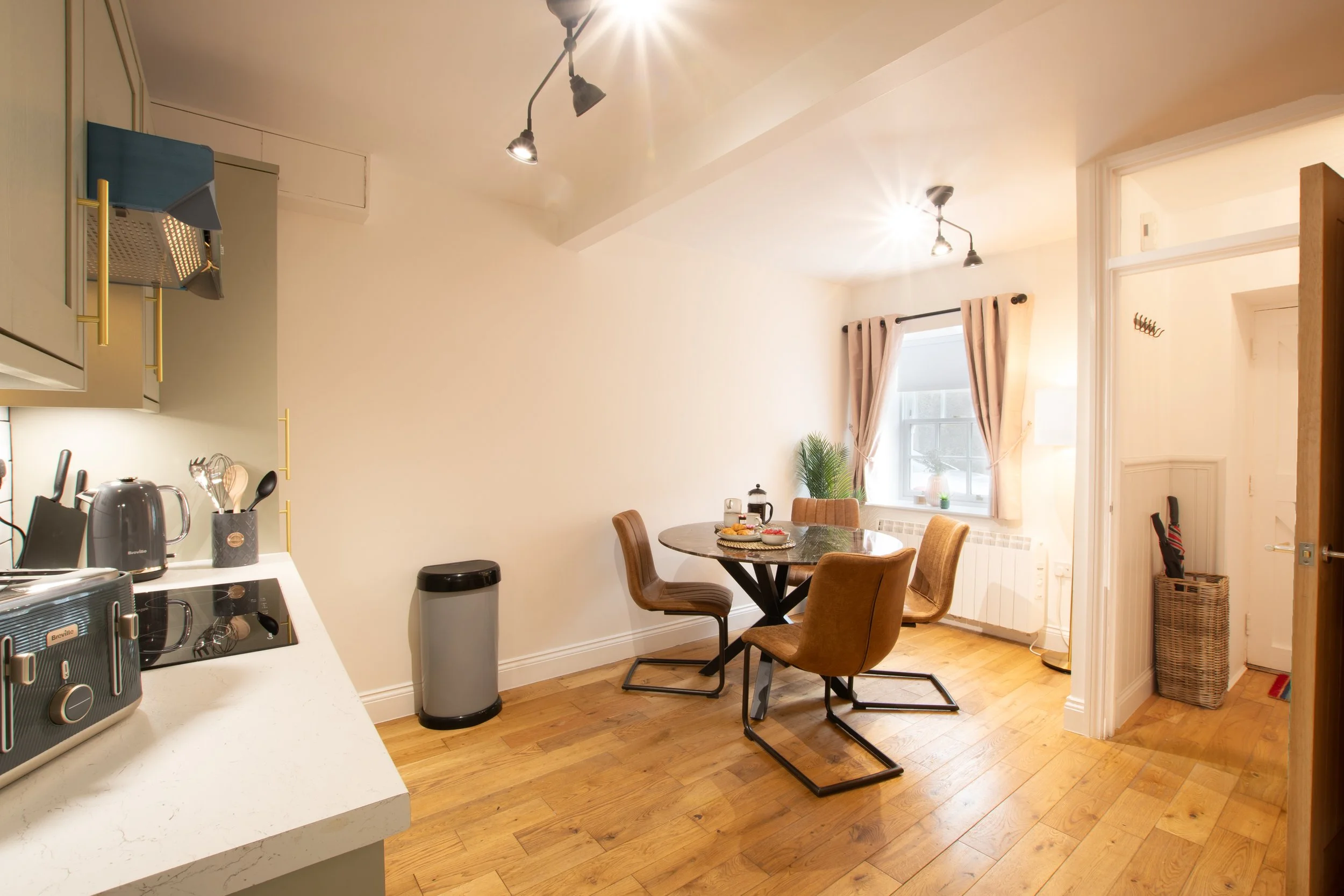 A cozy kitchen and dining area with a round wooden table, four brown chairs, a window with beige curtains, and a wall-mounted coat rack. The kitchen has white cabinets, a countertop with small appliances, and ceiling track lighting. There is hardwood