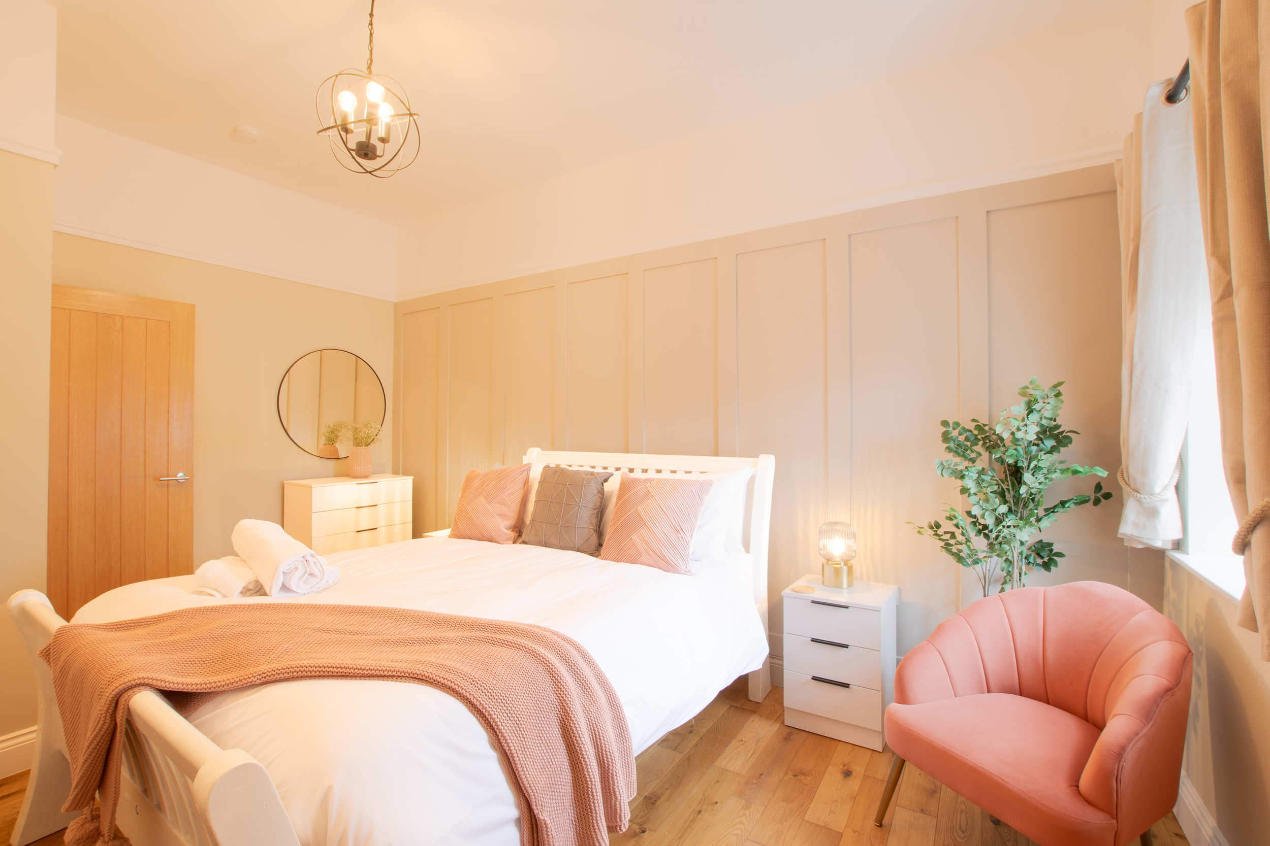 A bedroom with a white bed, pink and brown pillows, a pink armchair, a white nightstand with a lamp, a window with curtains, a plant, a dresser with a round mirror, and a ceiling light fixture.