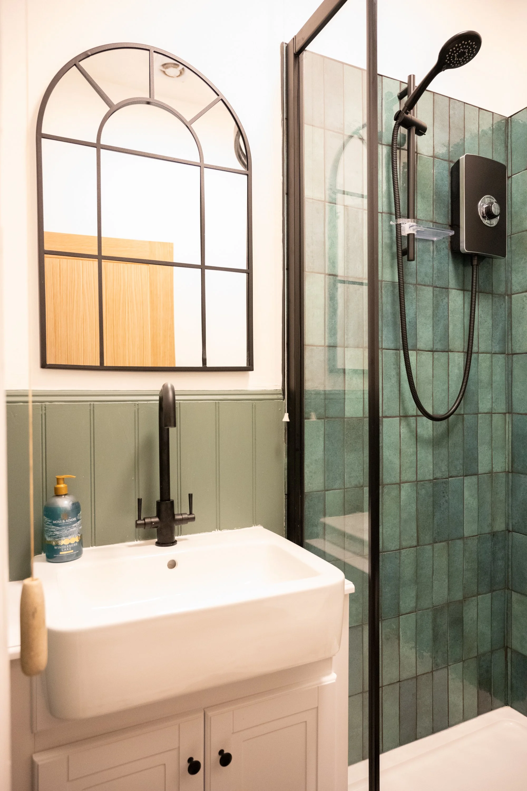 Bathroom with a white sink, a black faucet, a wooden mirror frame, a green cabinet wall, a soap dispenser, and a glass shower enclosure with green tiles and a mounted showerhead.