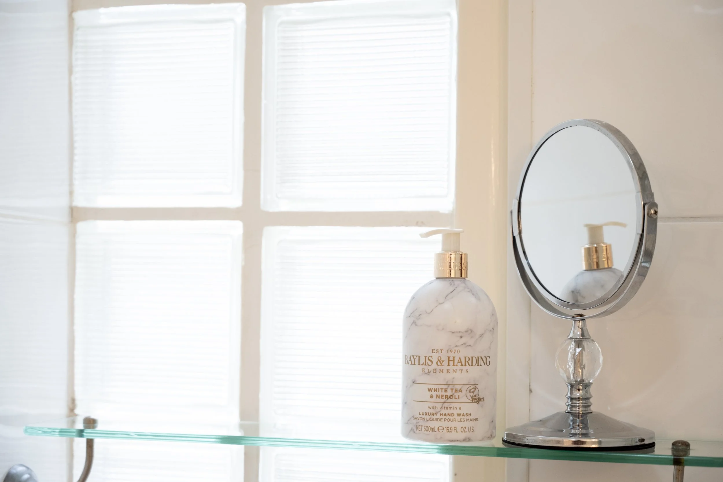 A white bottle of luxury hand wash labeled Baylis & Harding on a glass shelf. Behind it is a round mirror and a window with frosted glass panes that allow light to filter in.