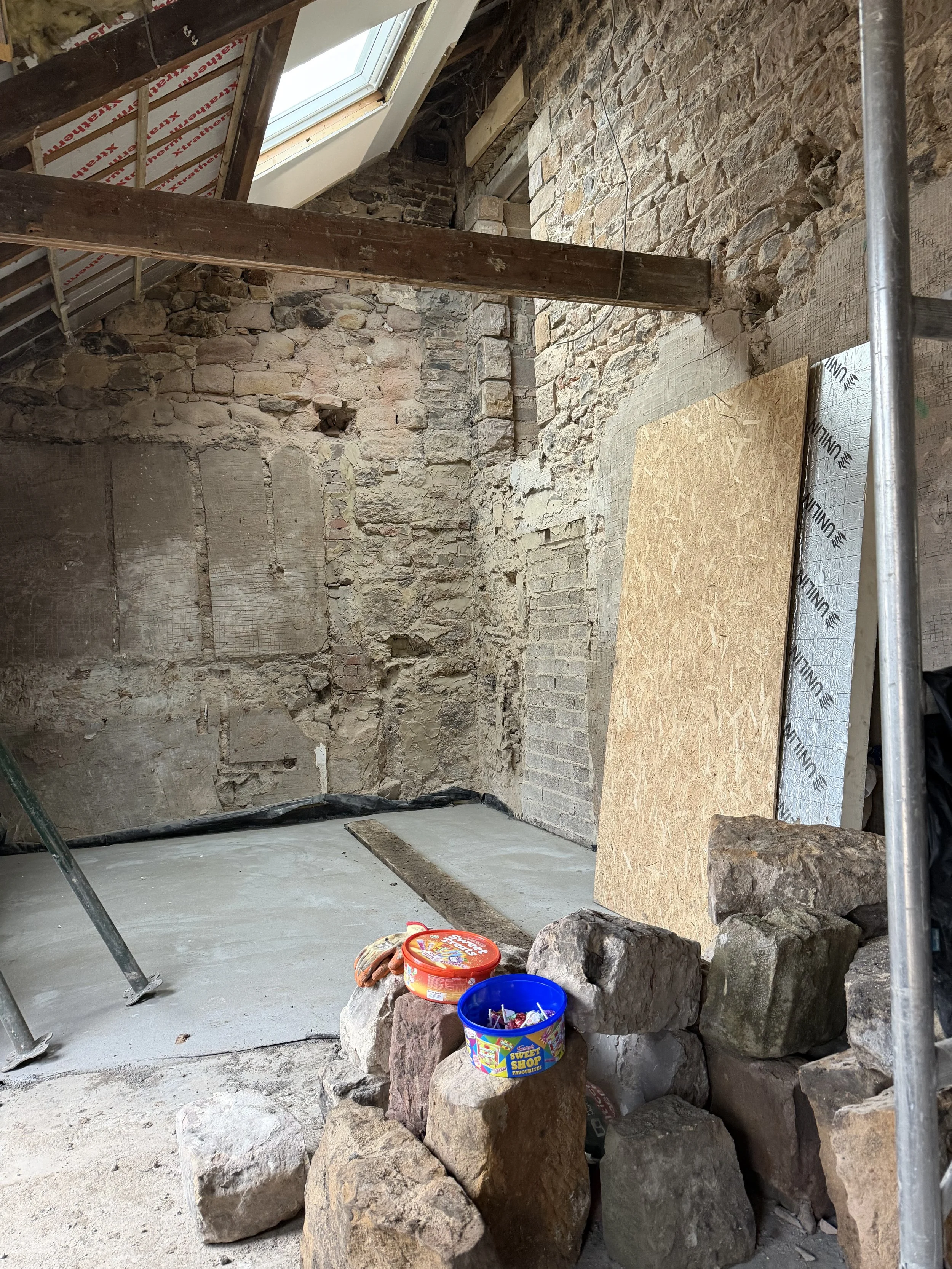 Unfinished attic with exposed stone walls, a skylight window, and construction materials including large rocks, plywood, and insulation. There are tools and buckets, indicating ongoing renovation work.
