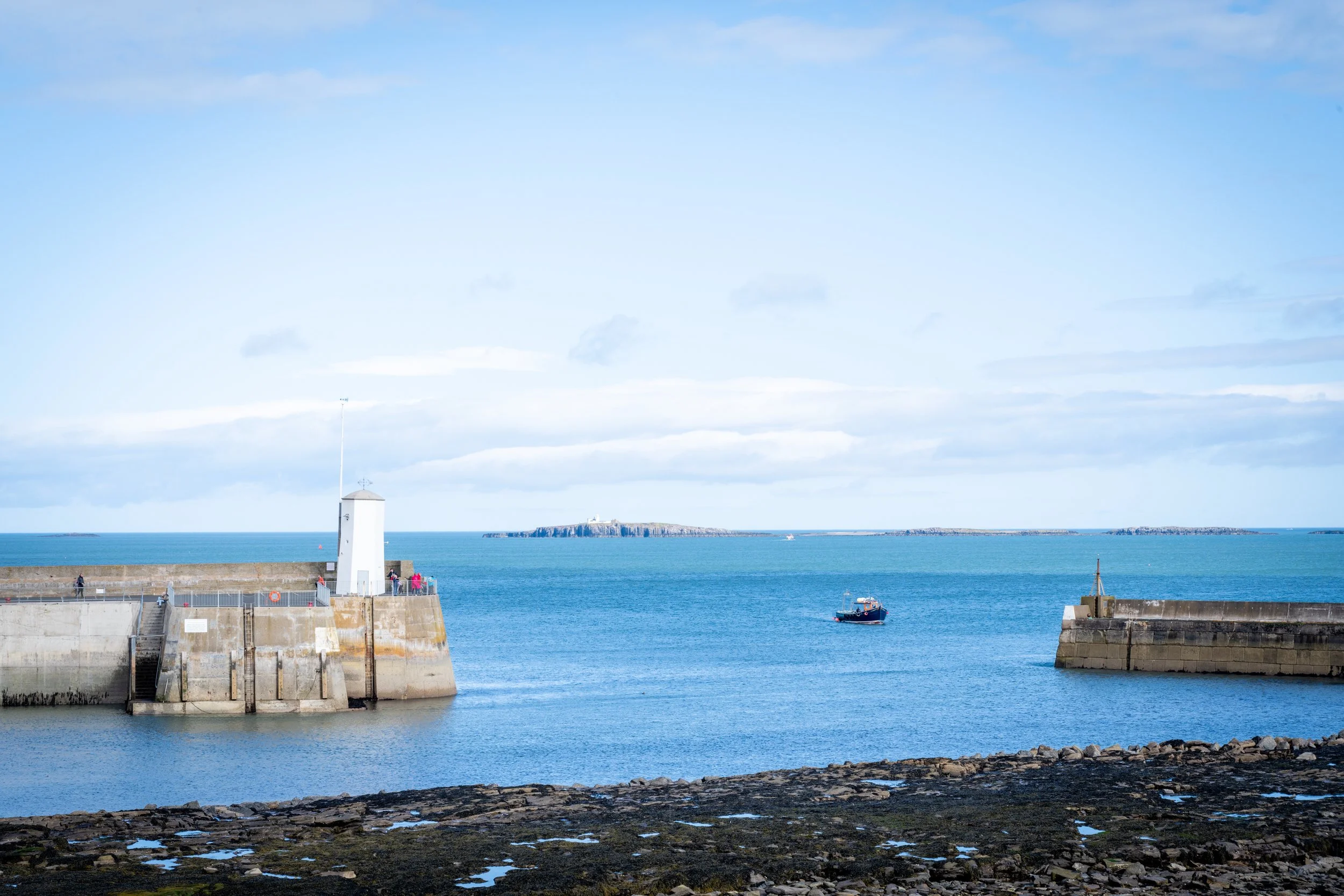A harbor scene with a lighthouse on a concrete pier, a boat in the water, and a distant island under a partly cloudy sky.