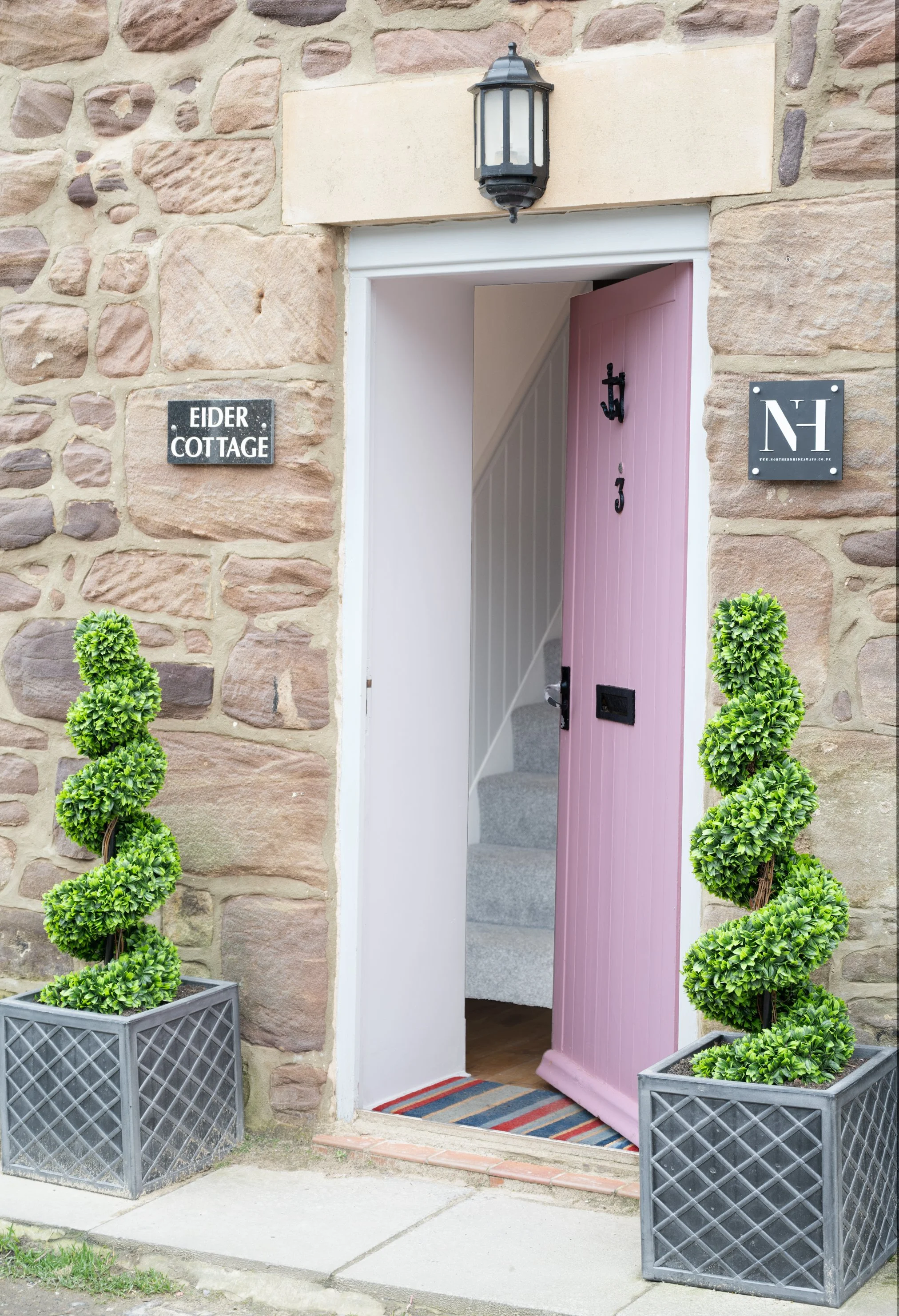 Open doorway to a stone cottage with a pink door partially open. Two black planters with shaped bushes are on either side of the doorway, and a lantern is mounted above the door. Two black signs with white text are on the stone wall, reading 'Elder C