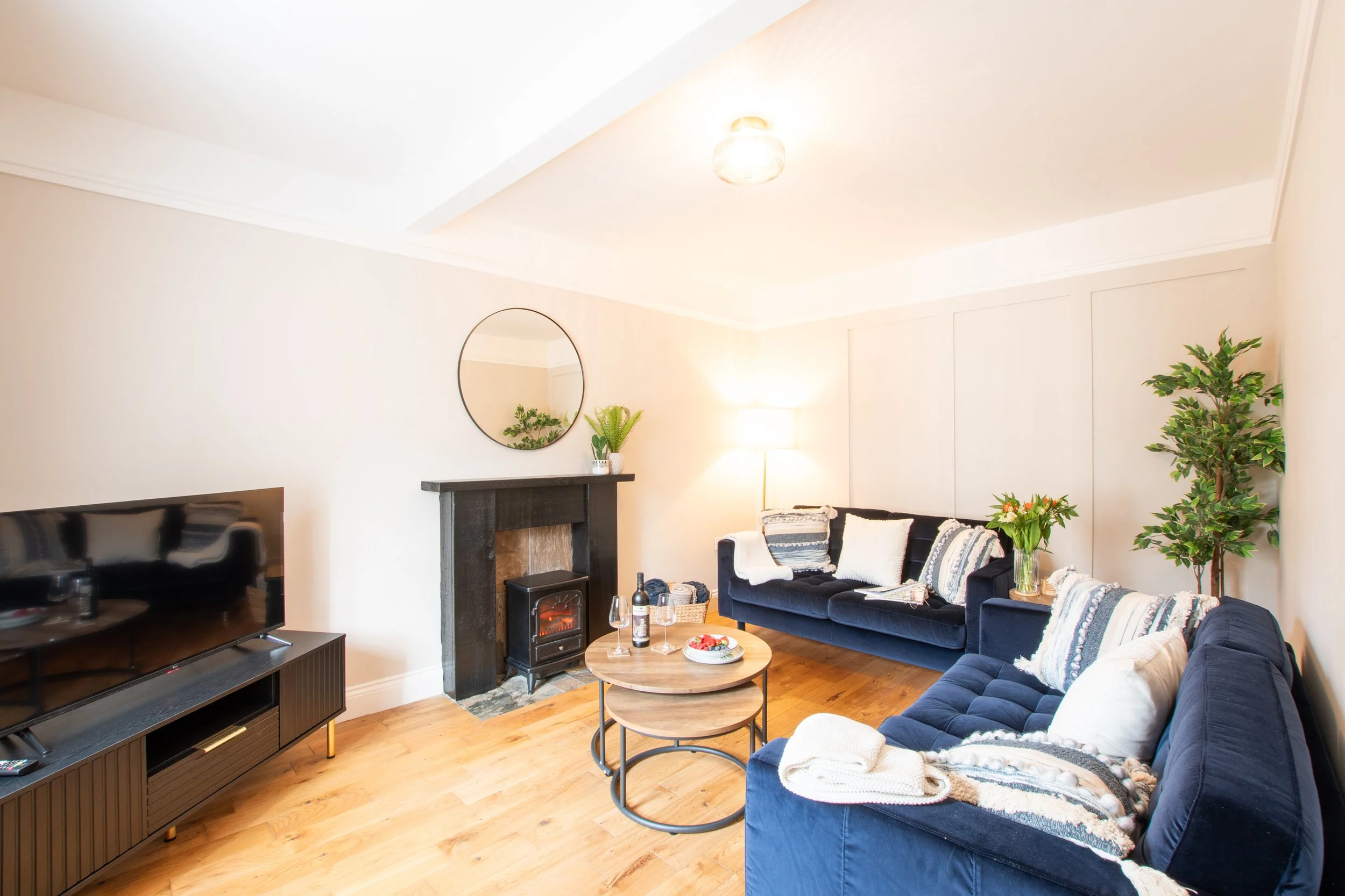 Living room with black sofas, a wooden coffee table with drinks and snacks, a flat-screen TV, a fireplace, plants, a mirror, and soft lighting.