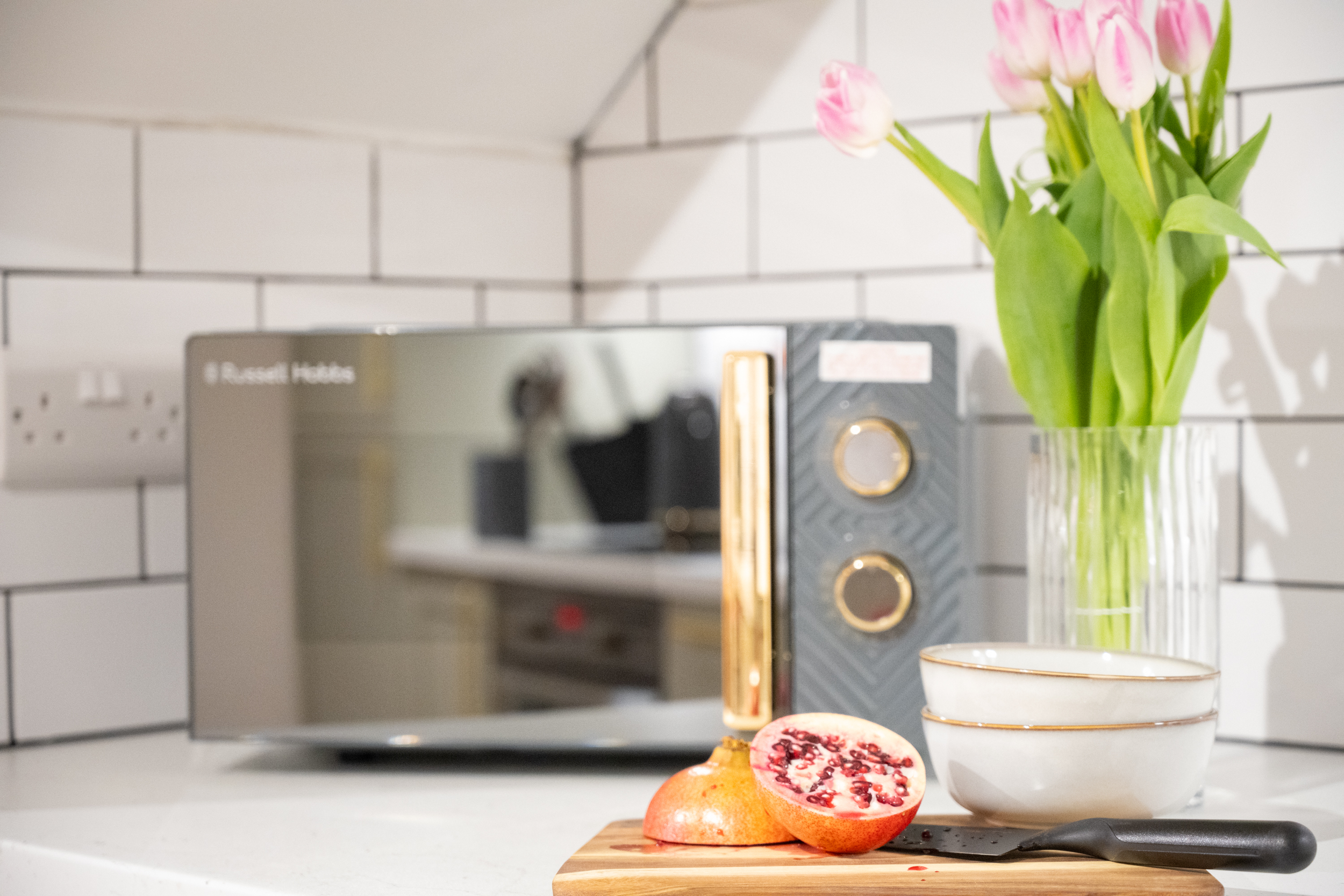 Kitchen countertop with sliced pomegranate, whole pomegranate, and a knife on a wooden cutting board, with a vase of pink tulips, a coffee machine, and stacked white bowls in the background.