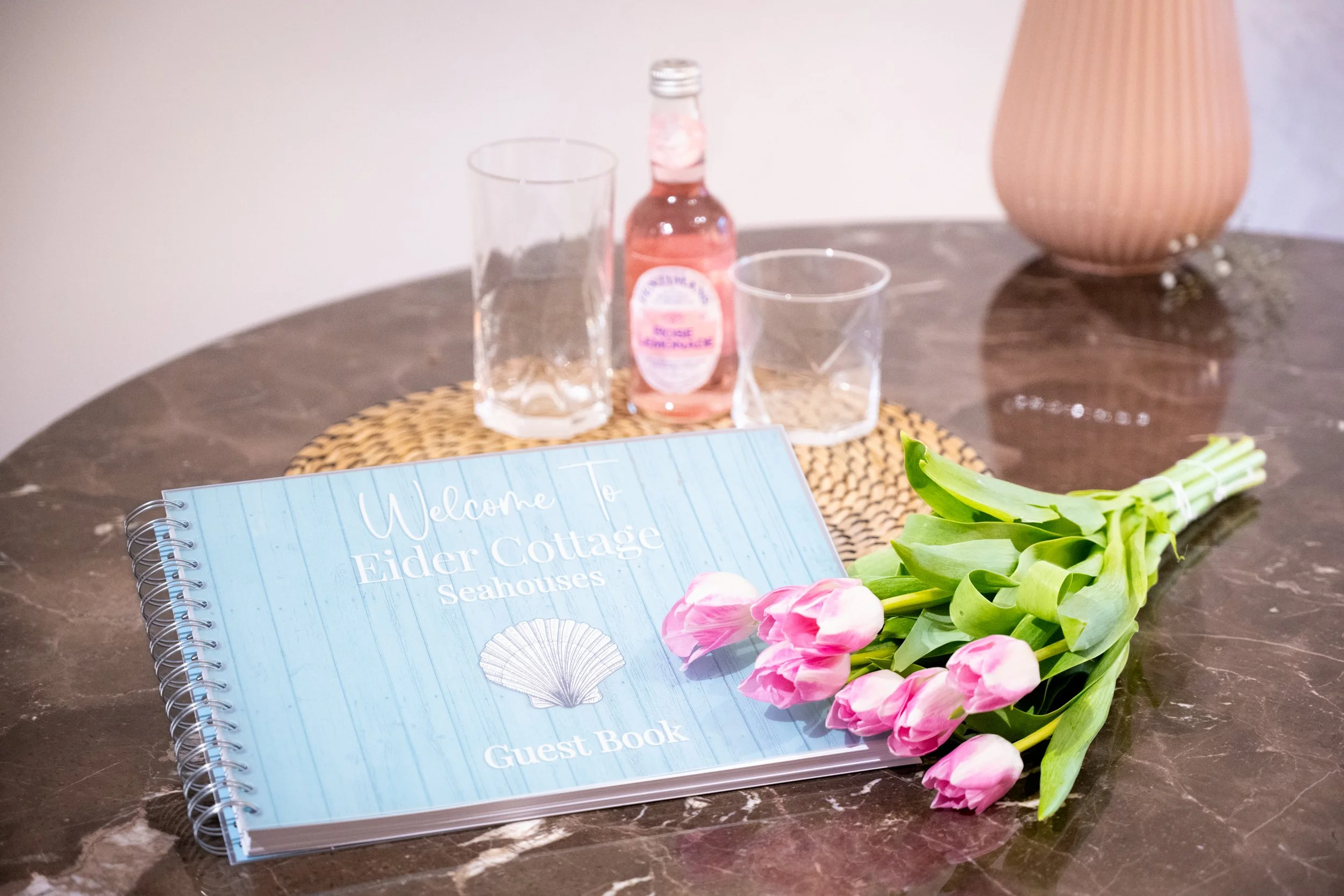 A guest book with a blue cover and white text reading 'Welcome To Eider Cottage Seahouses,' a bouquet of pink tulips, a glass bottle of pink lotion, and two empty glasses on a woven placemat on a dark marble table with a pink textured vase in the bac