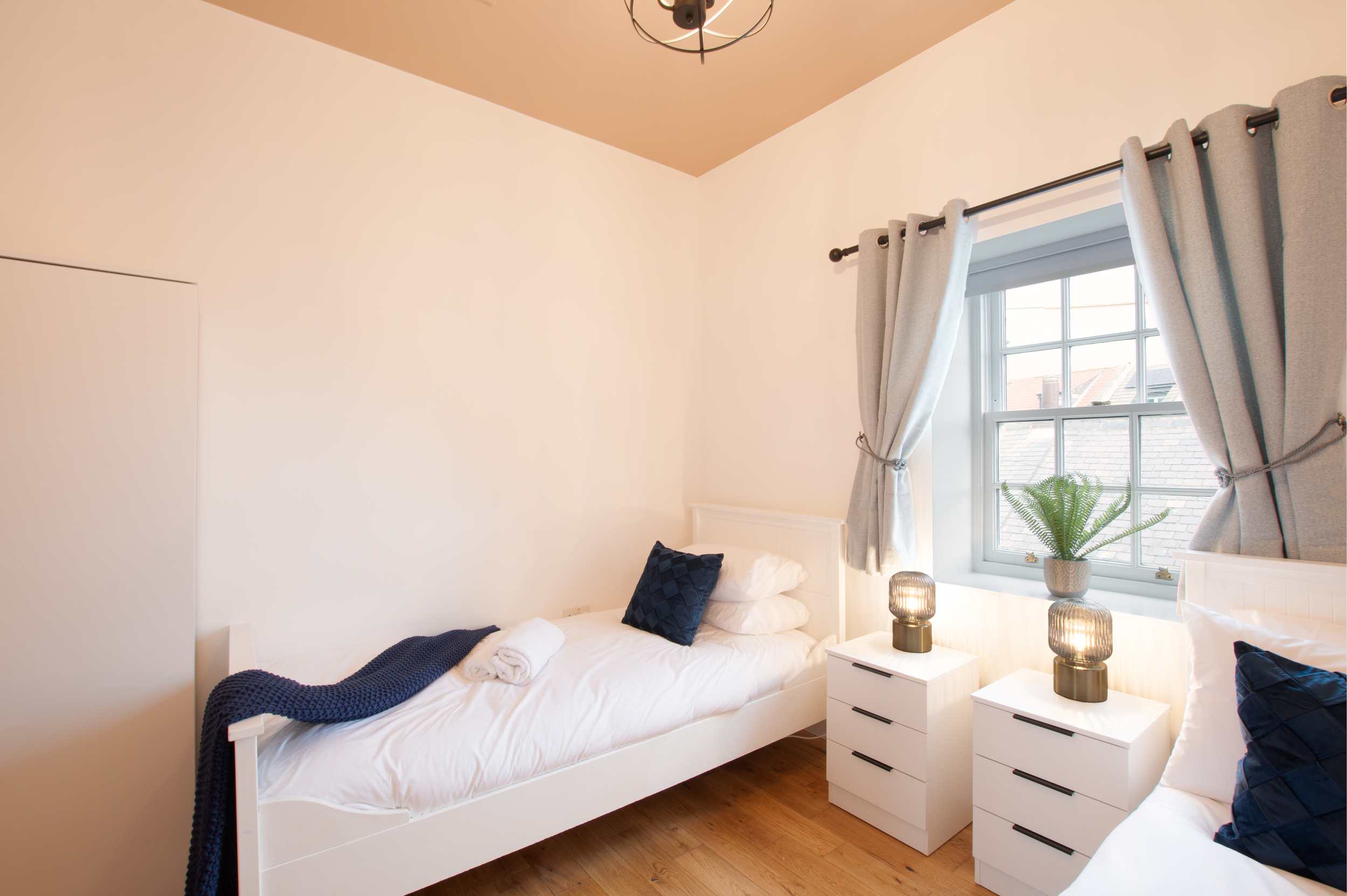 A tidy bedroom with a white bed, dark blue pillows, a dark blue blanket, and a towel, next to a white nightstand with two lantern-style lamps, a window with light gray curtains, and a potted fern.