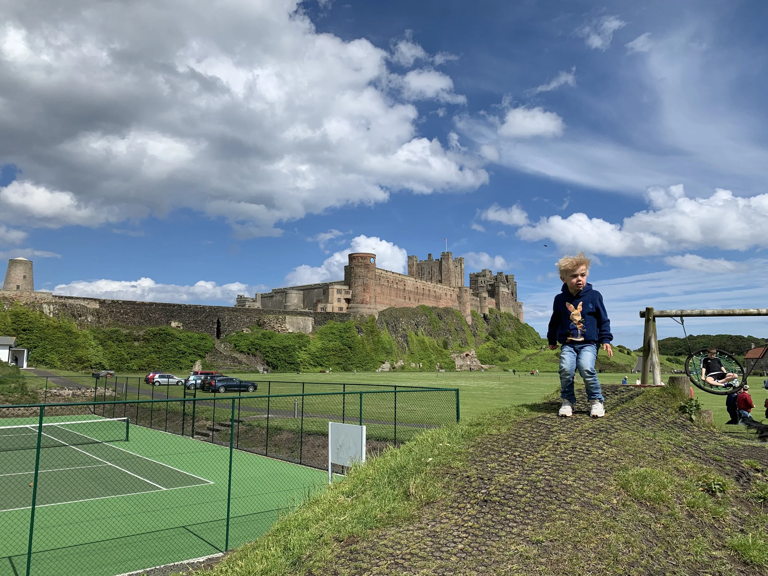 A young boy with blond hair wearing a navy hoodie and jeans standing on a playground in front of a historic castle on a partly cloudy day.