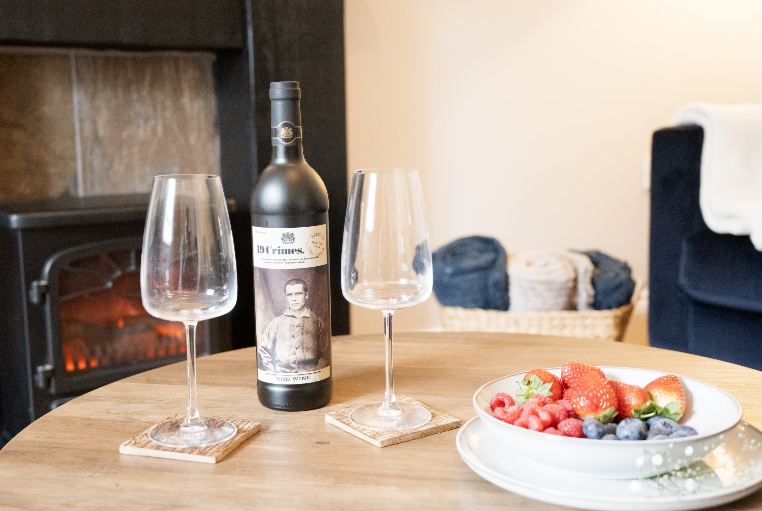 A bottle of red wine on a wooden table with two empty wine glasses, a plate of mixed berries, and a fireplace in the background.