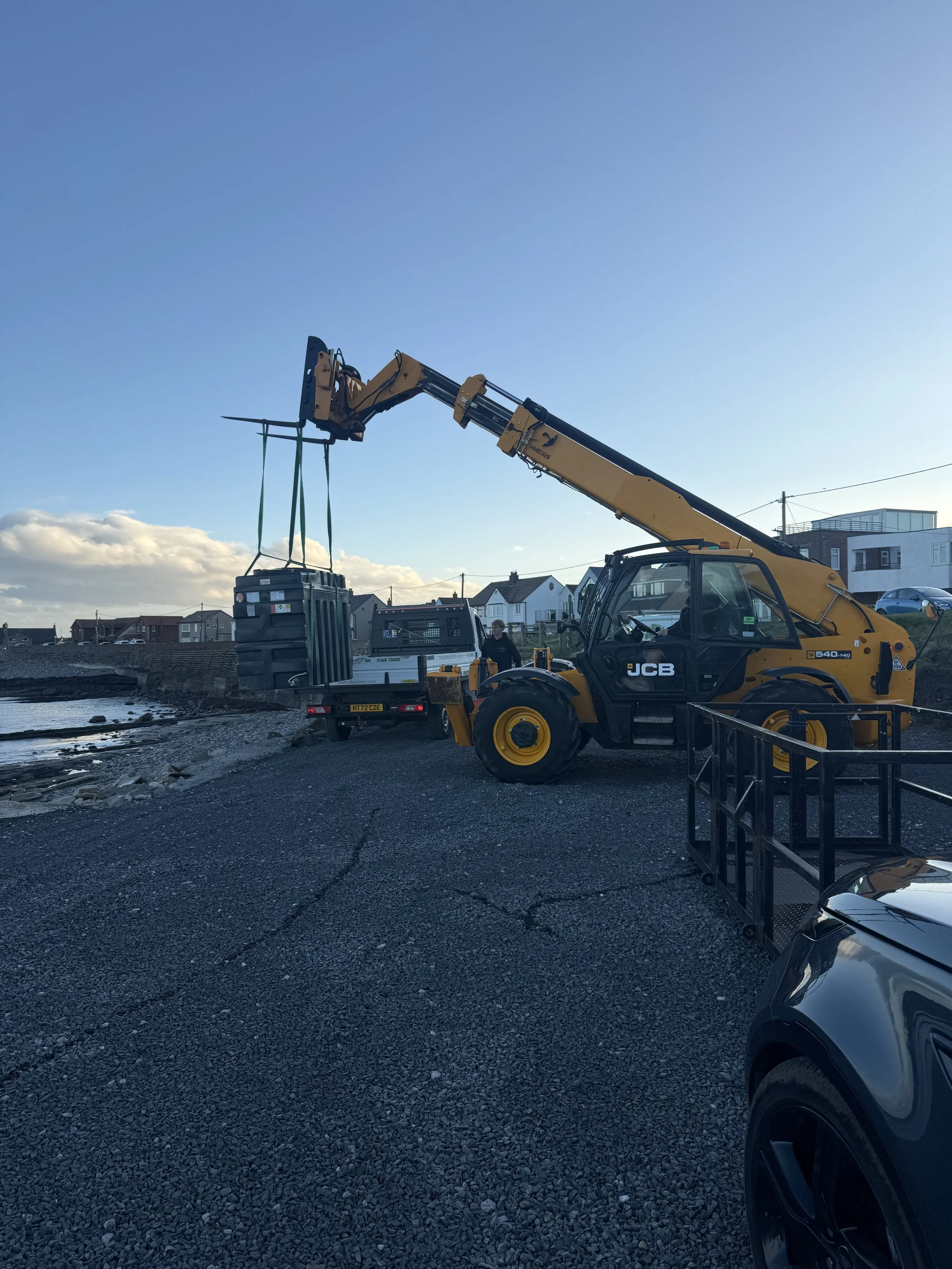 A yellow JCB telescopic handler lifting a black trash bin with green straps, parked on a gravel surface along a waterfront with houses and a blue sky in the background.
