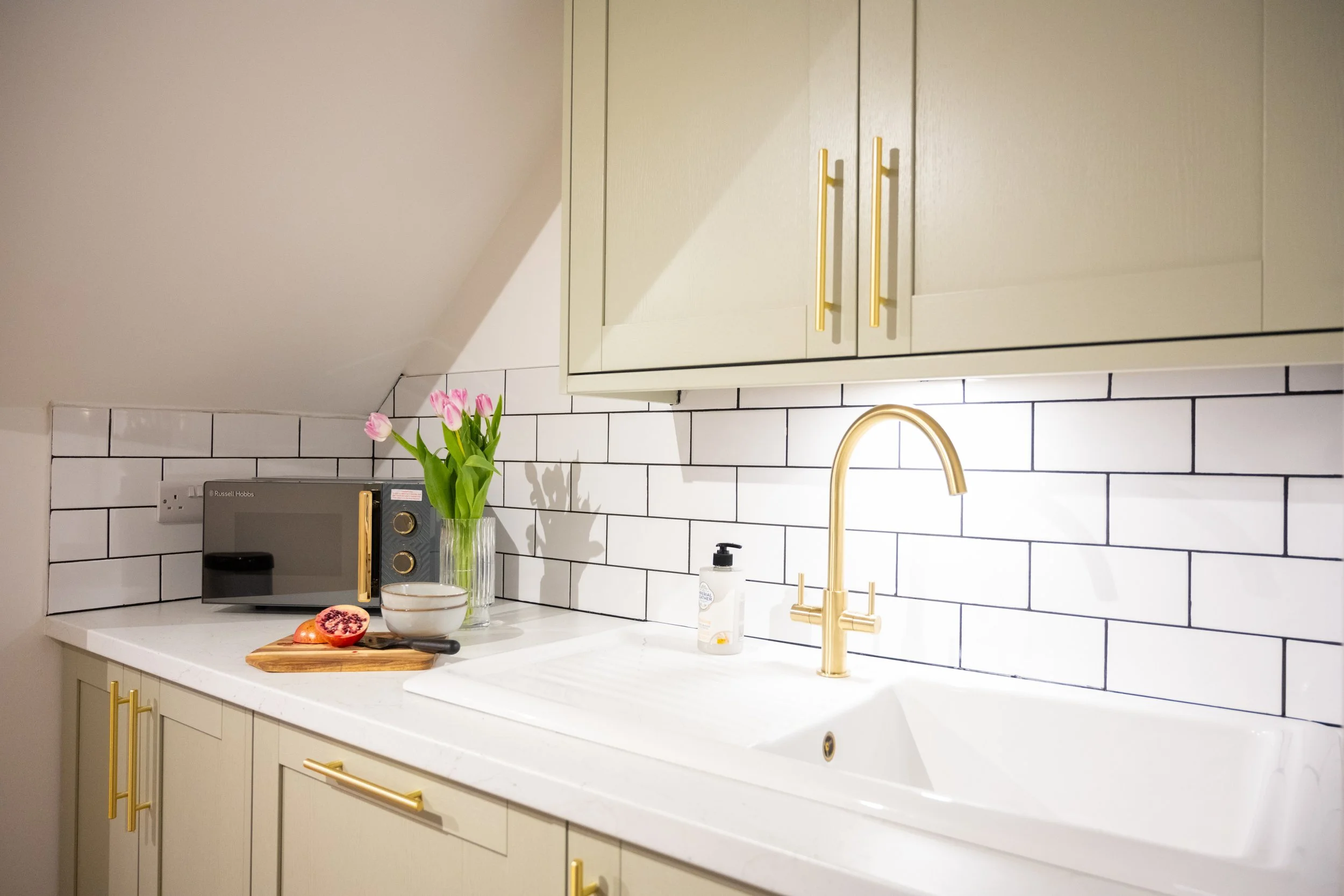 A kitchen workspace with white countertops and a black and white subway tile backsplash. There are gold cabinet handles, a gold faucet, a black microwave, a vase with pink tulips, a halved pomegranate, a bowl, a knife, and a soap dispenser on the cou