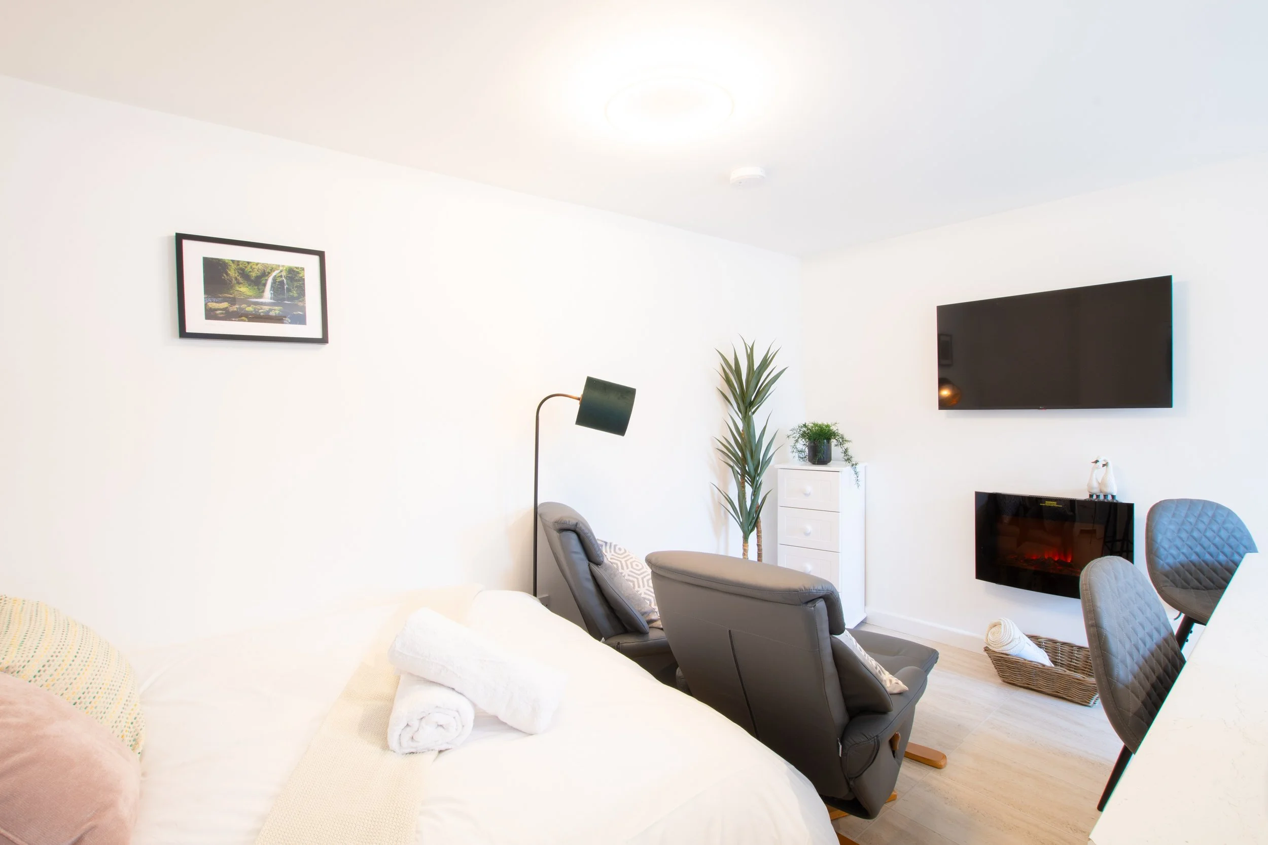 Living room with white walls, a black flat-screen TV mounted on the wall, a tall green plant, a small white cabinet, framed picture of a waterfall, a floor lamp, and chairs with rolled towels.