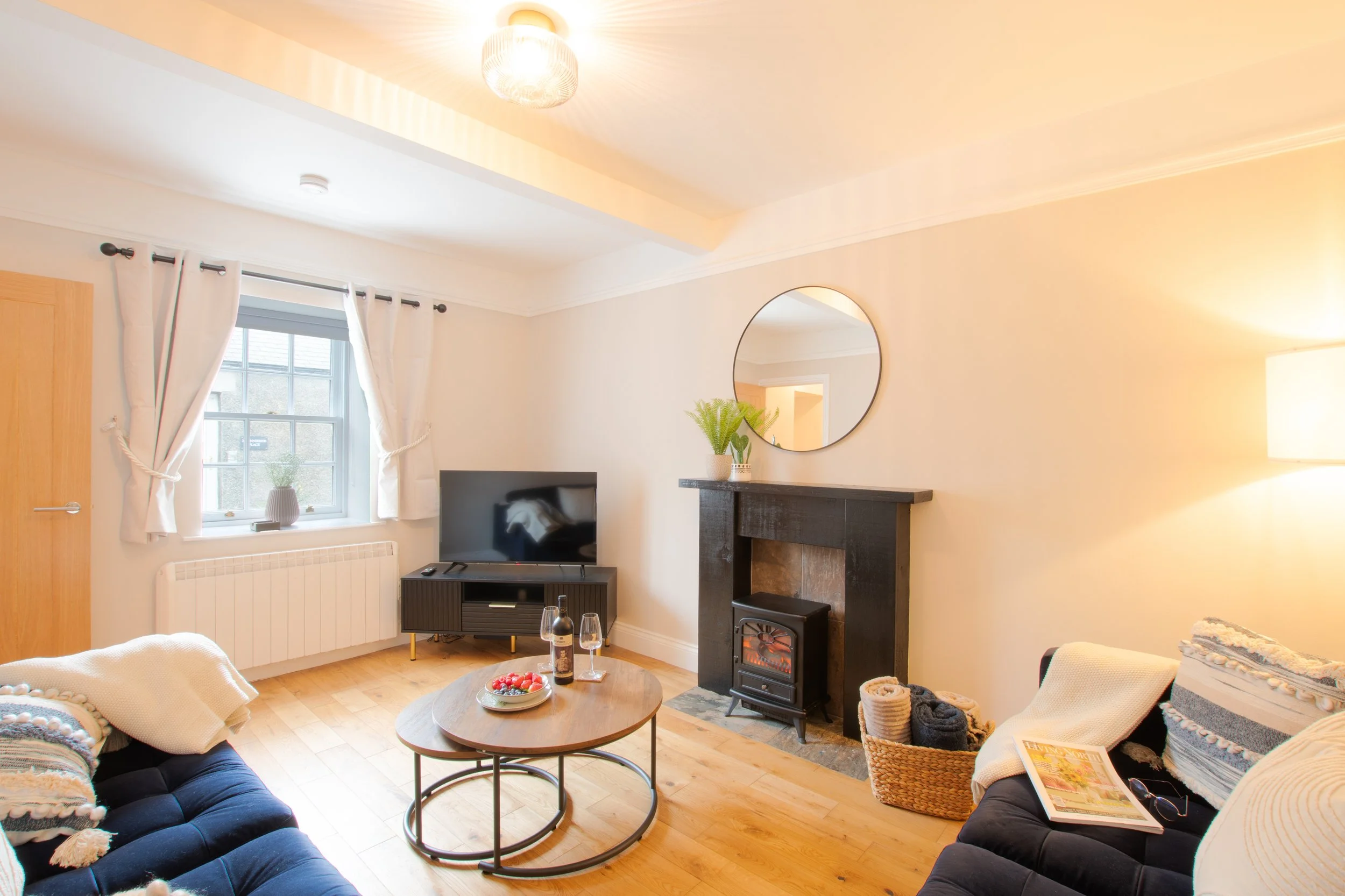 Living room with white walls and wooden floors, featuring a window with white curtains, a black fireplace, a flat-screen TV, a round coffee table with a bottle of wine and a bowl of berries, a blue sofa with a blanket, a basket with rolled towels, an