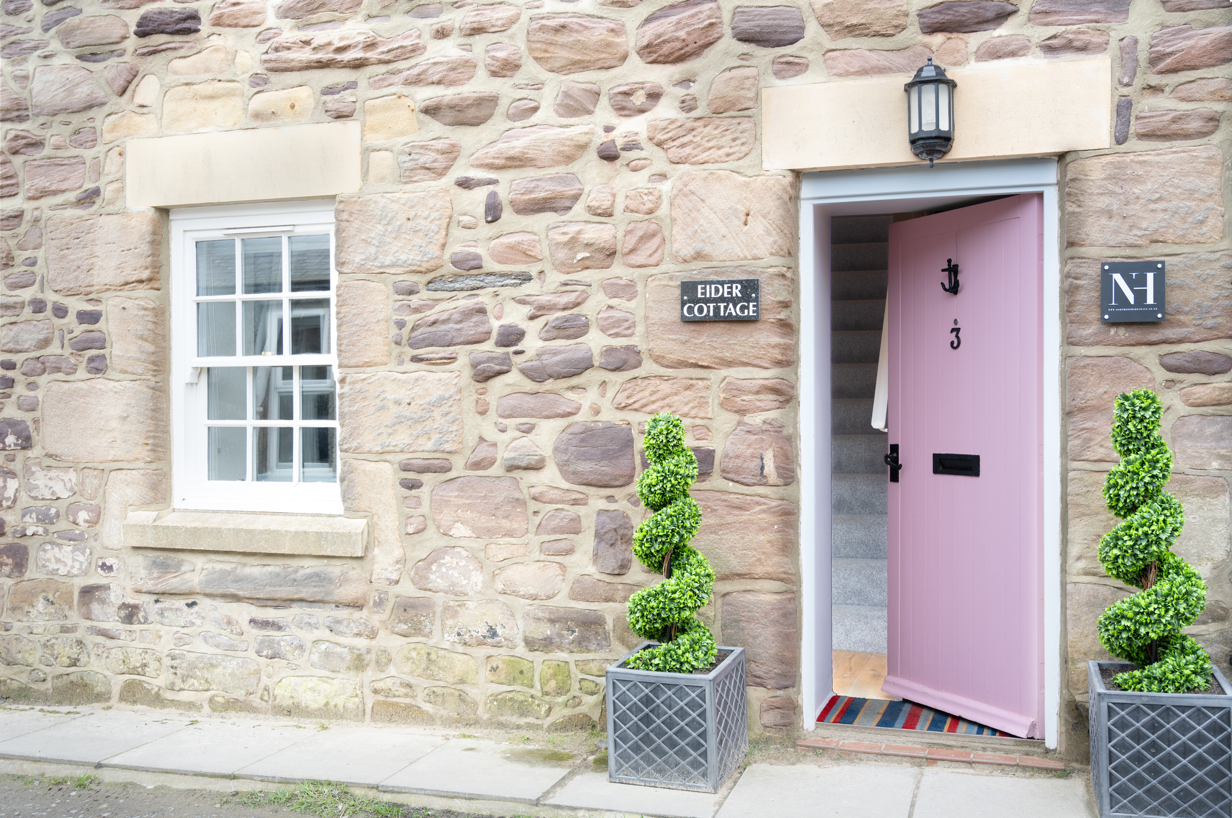 The front of a stone house called Eider Cottage with a pink front door, greenery in square planters, and a black outdoor light fixture above the door.