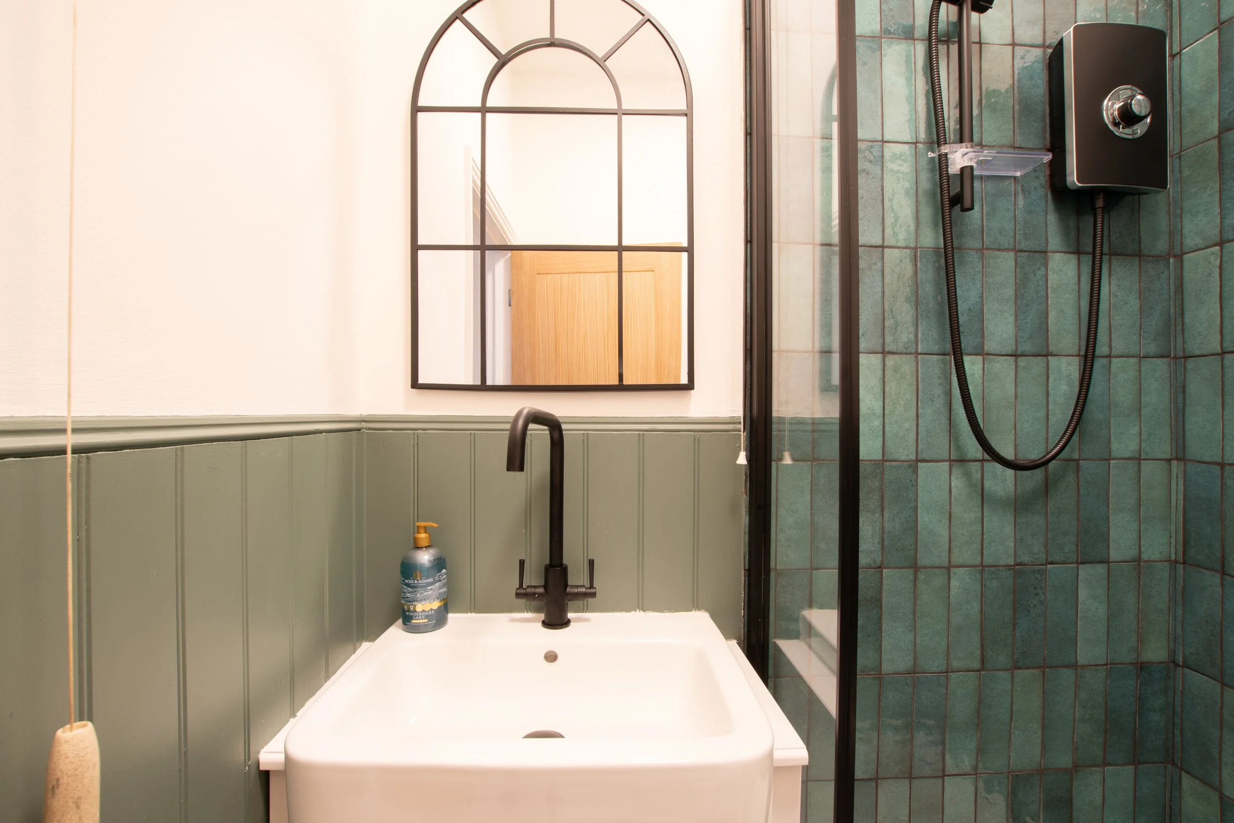 Bathroom with a shower area on the right, featuring green tiles and glass door. In the foreground, a white sink with a black faucet, and a soap bottle on the counter. A mirror with a black frame hangs on the wall, reflecting a door and part of a room