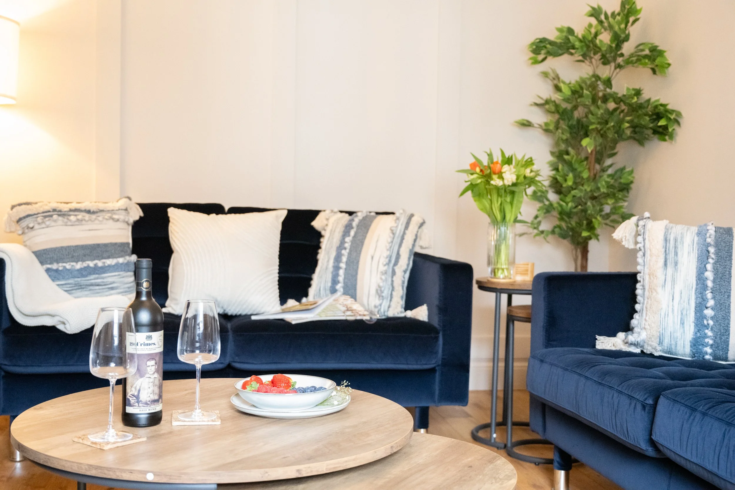 Living room with navy blue sofa, white and blue throw pillows, wooden coffee table with a bottle of red wine and glasses, and a bowl of strawberries and blueberries. A side table with a vase of white and orange flowers, a tall green houseplant, and a