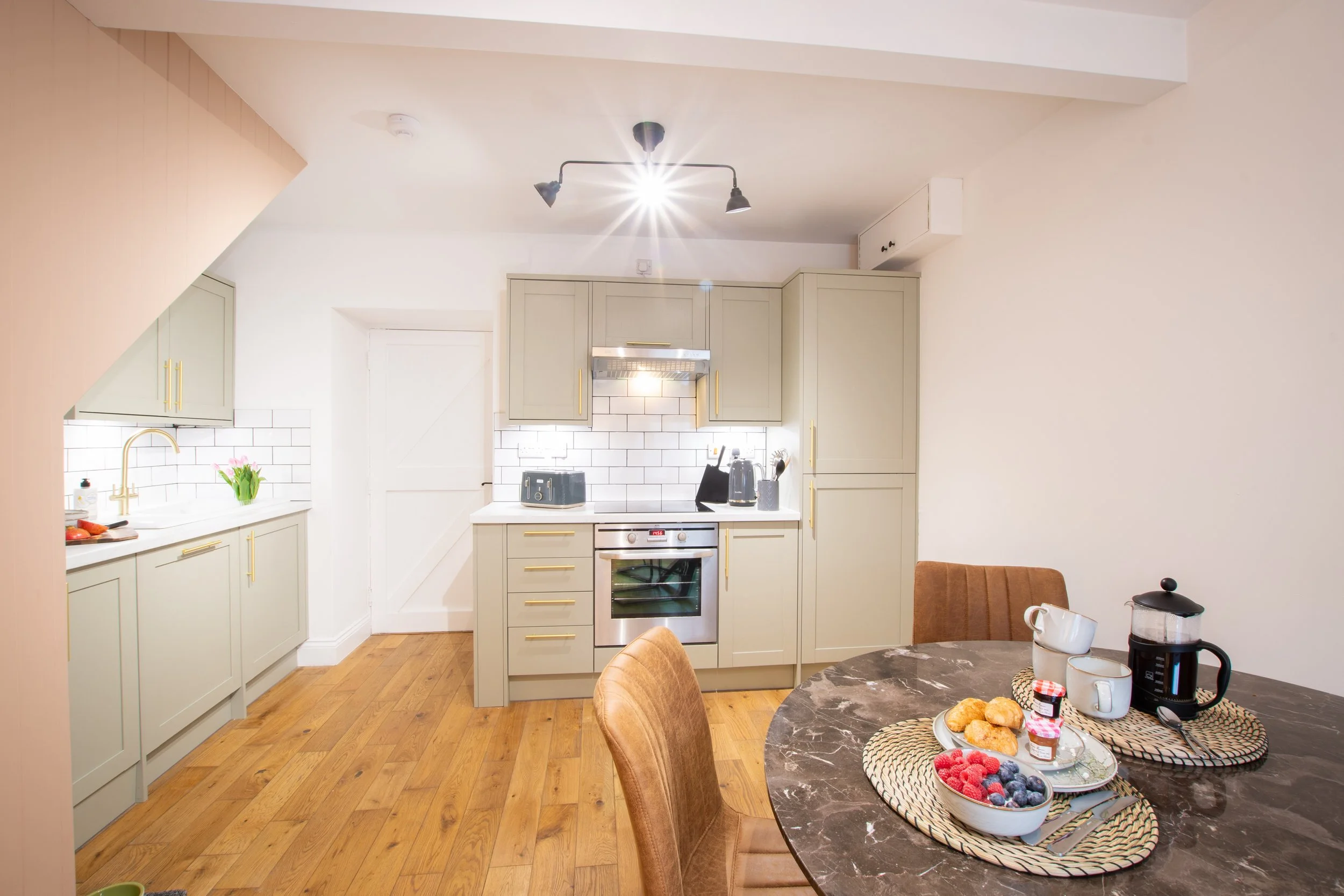 A kitchen with beige cabinets, a white subway tile backsplash, a built-in oven, and a marble dining table with breakfast foods and coffee mugs.