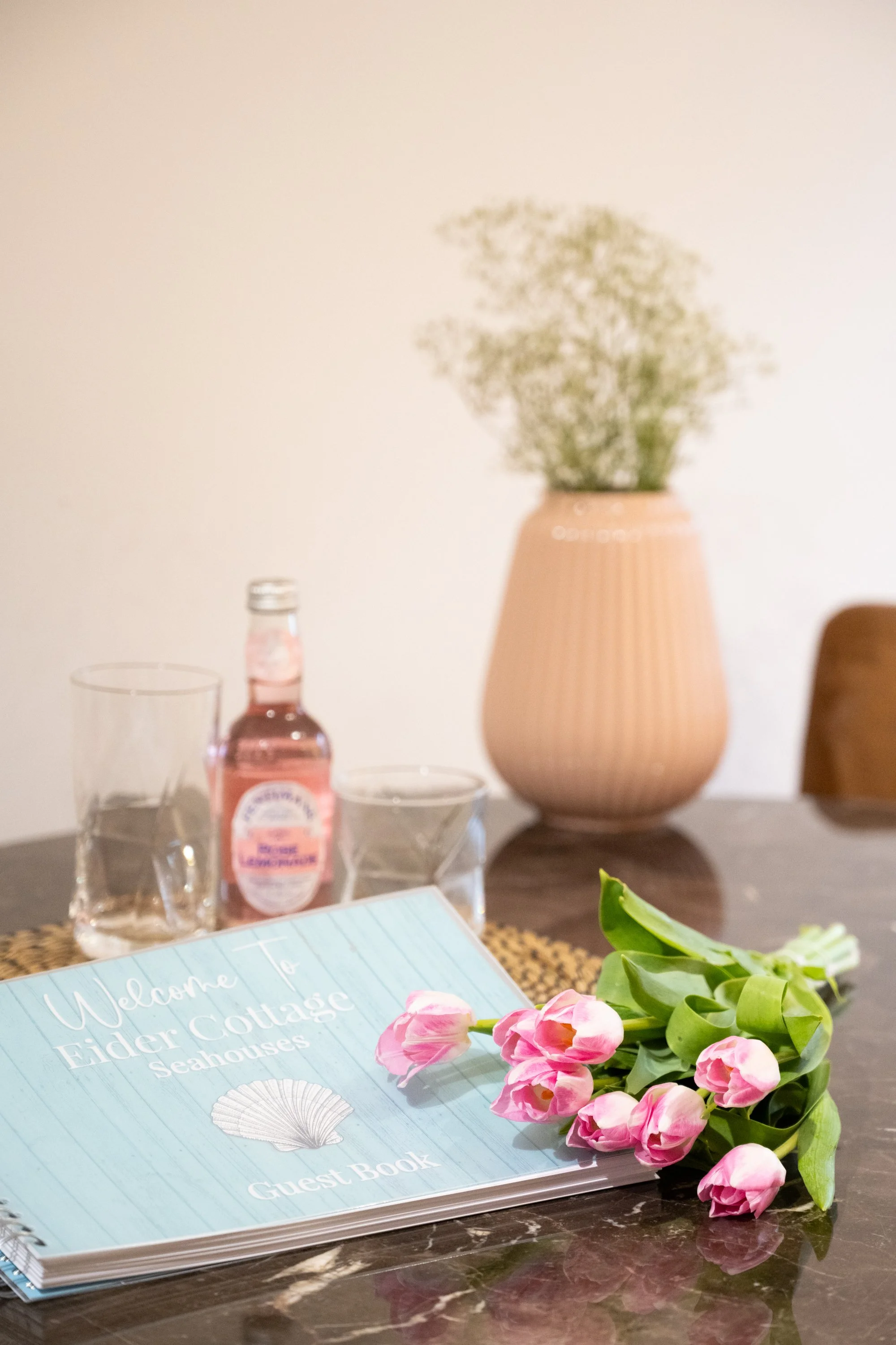 A table featuring a light blue guest book titled 'Welcome to Tiger Cottage Elder Seashells,' a bouquet of pink tulips, a bottle of pink soda, and two empty glasses, with a large beige vase with green plant in the background.