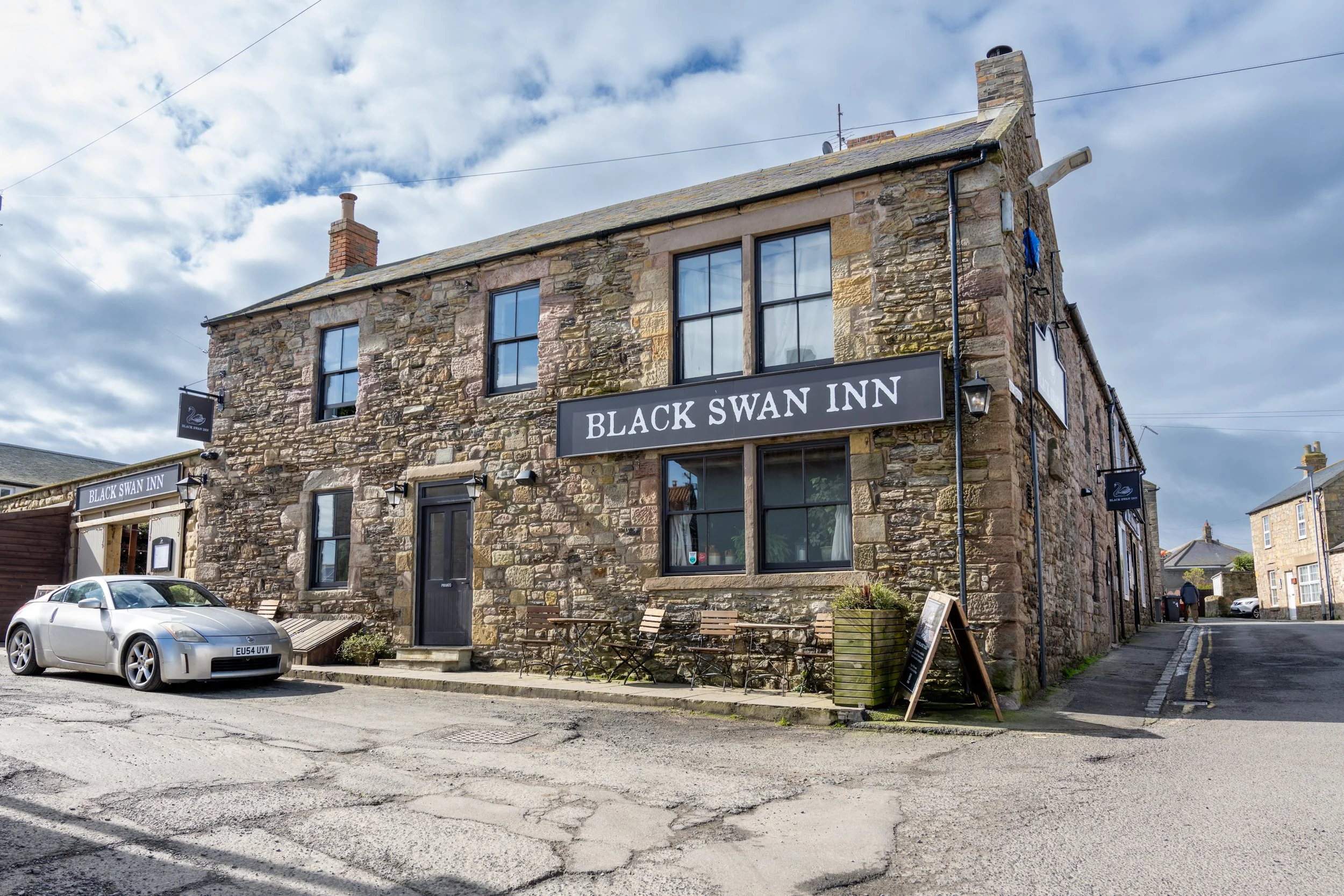 A rustic stone building with a sign reading 'Black Swan Inn.' There are a few cars parked in front, benches and planters near the entrance, and cloudy skies overhead.