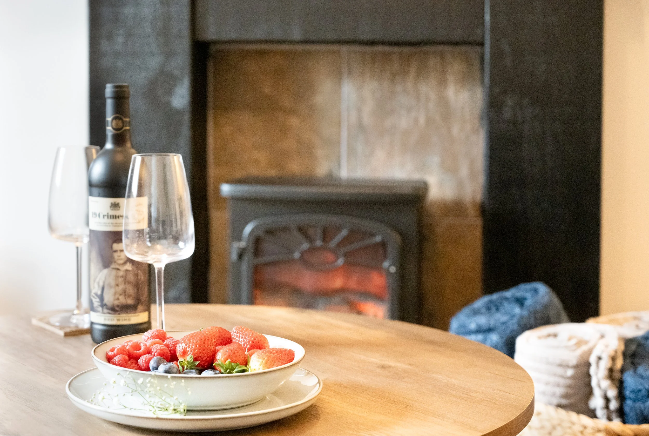 A table with a bowl of strawberries, raspberries, blueberries, and a strawberry plant, with a bottle of wine and two empty wine glasses nearby. In the background, a small stove or fireplace with a fire burning, and a fireplace mantel against a dark w