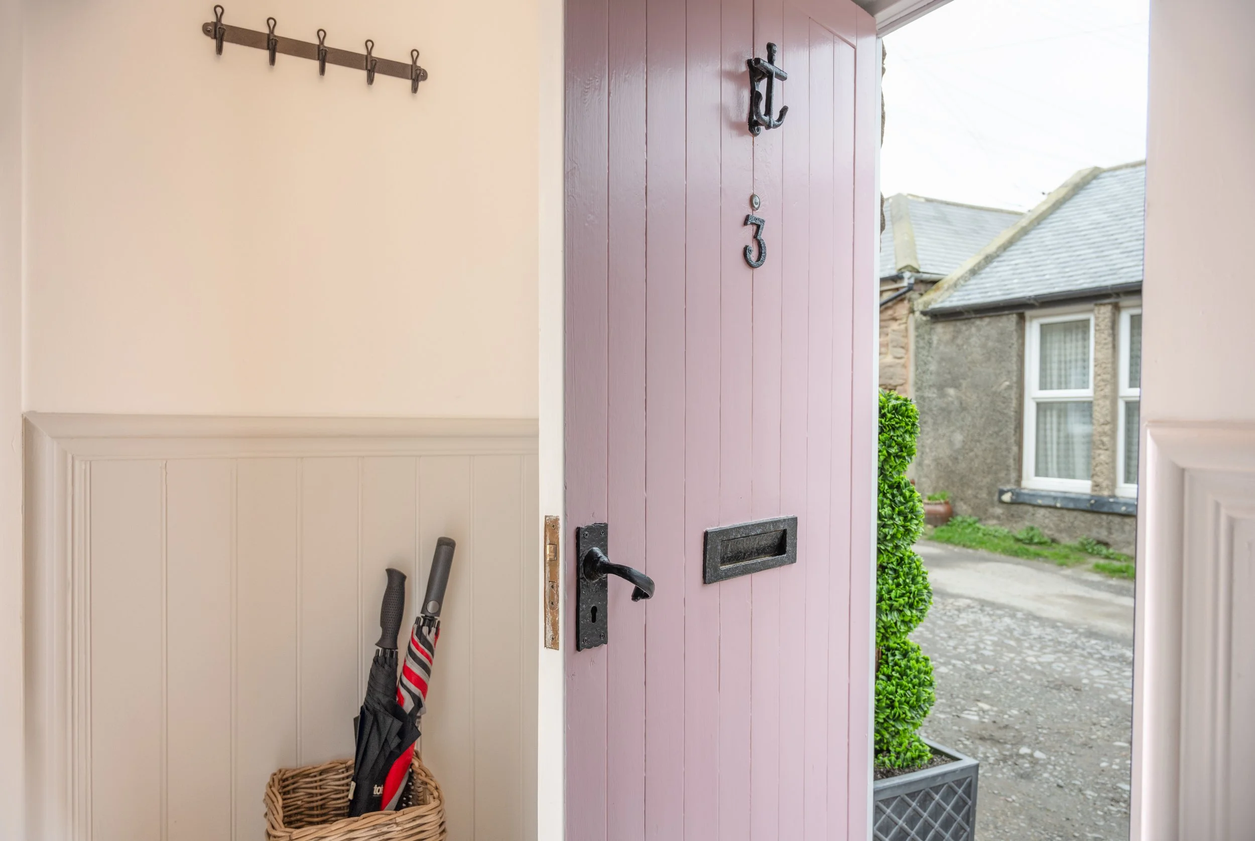 Open pink front door leading outside to a street with neighboring houses. A basket with umbrellas and a rake inside is on the left, and a coat rack with hooks is on the wall above.