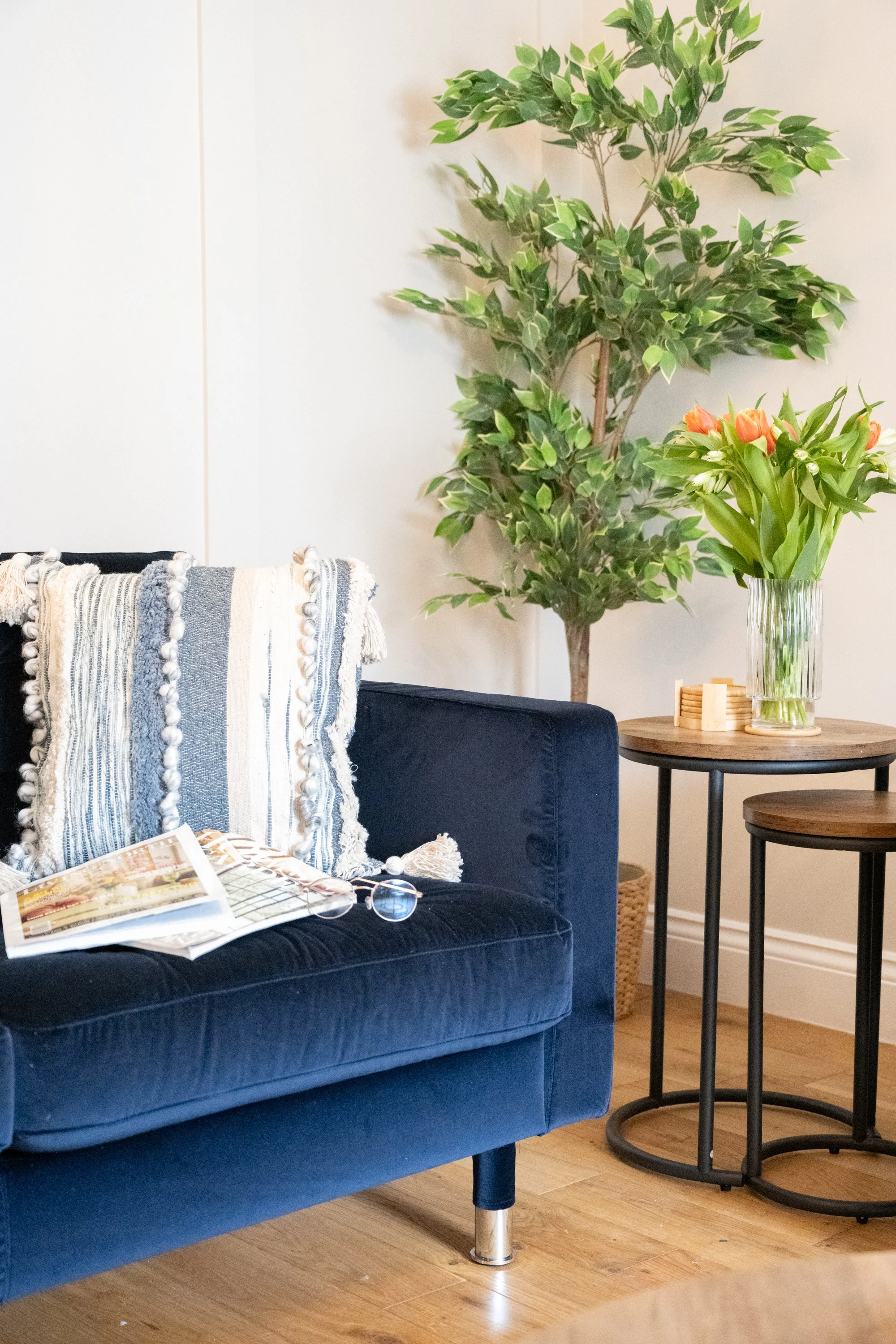 A living room corner with a navy blue velvet sofa, a striped and tasseled throw blanket, an open magazine, and a pair of glasses on the sofa. Beside the sofa, a round wooden side table holds a tall glass vase with orange tulips and white lilies. In t