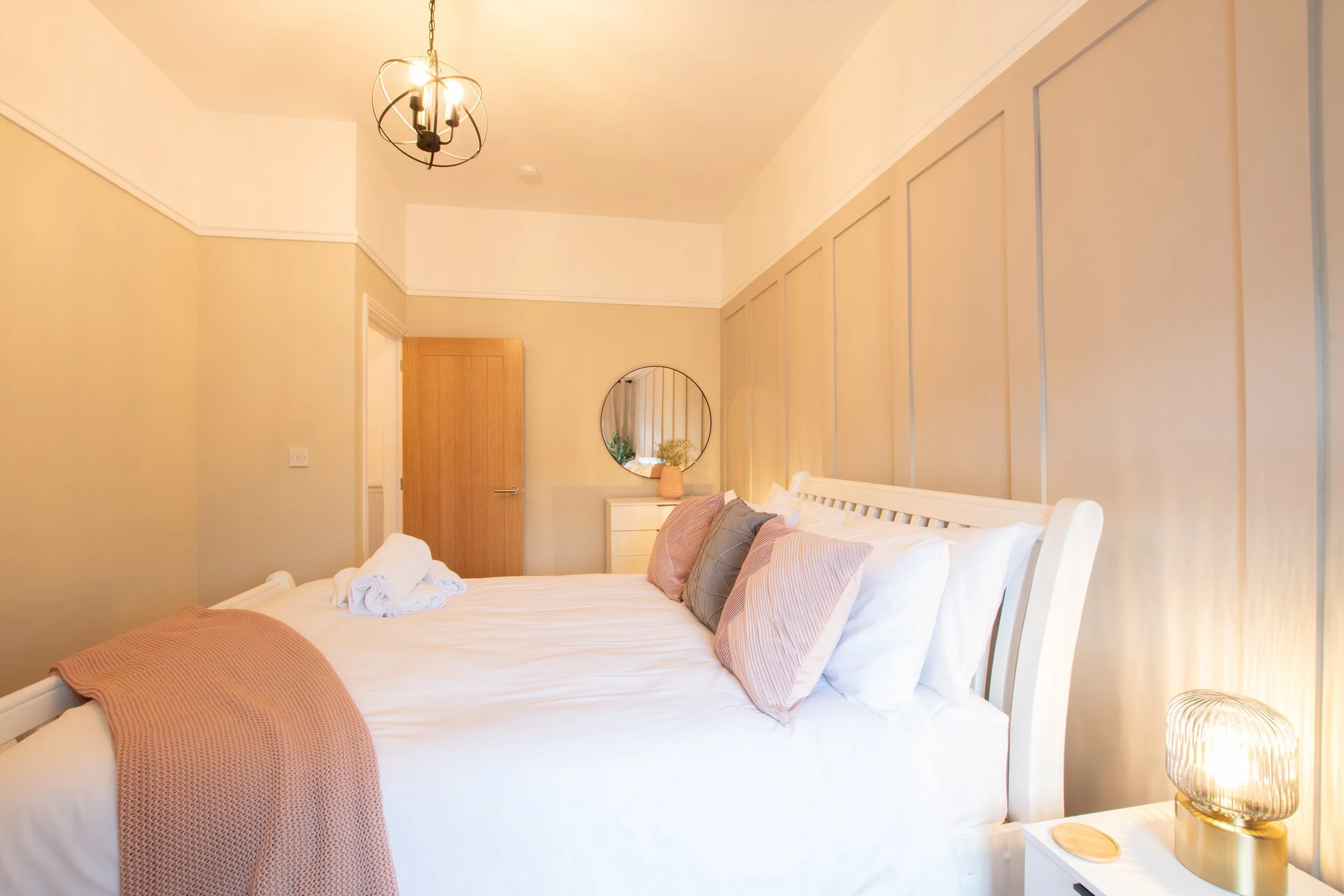 A cozy bedroom with a white bed, pink and gray pillows, and a pink throw blanket. A wooden bedside table has a lamp with a textured gold base and a round mirror hangs above a small white dresser decorated with a vase of flowers. The ceiling features 