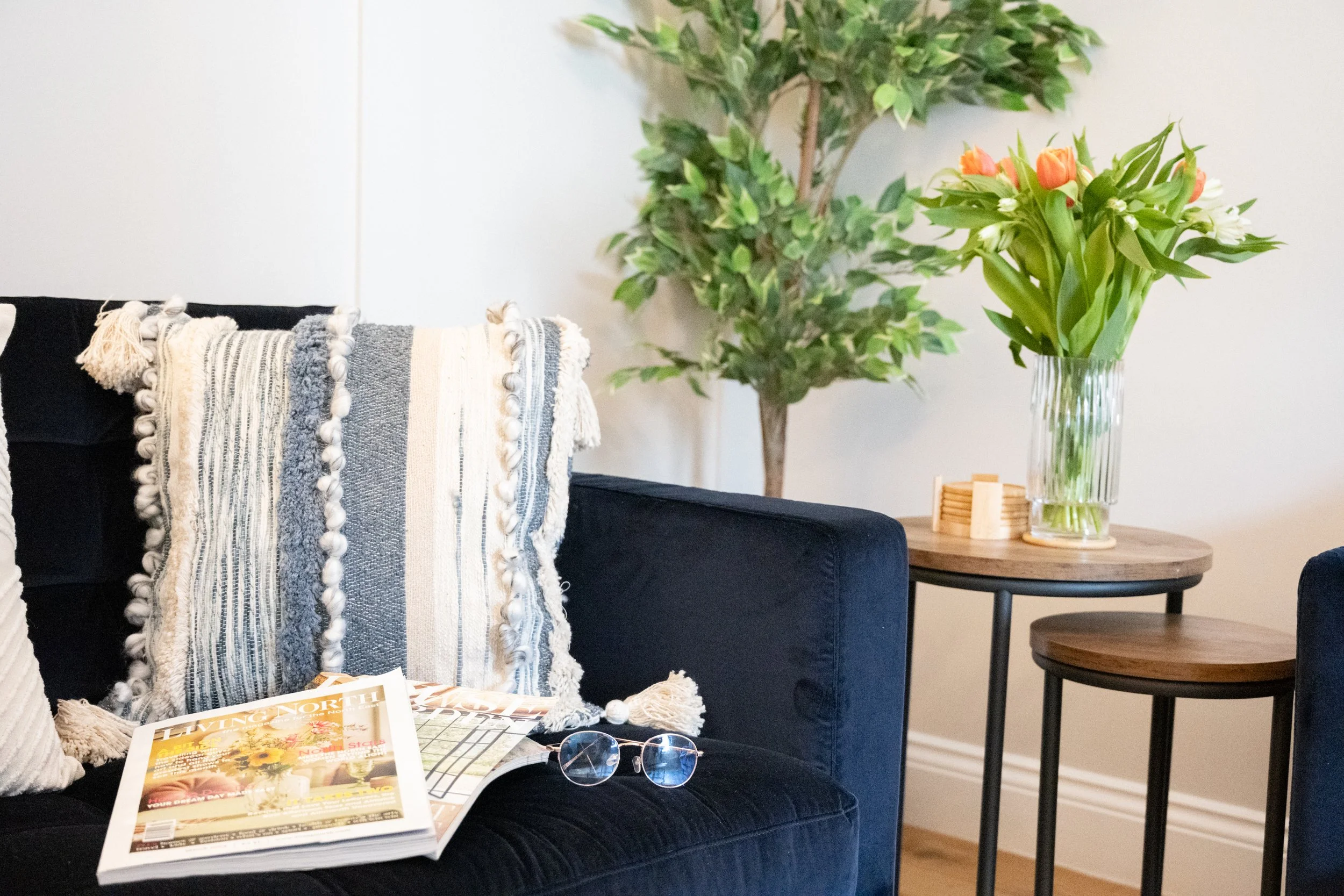 A living room scene featuring a navy blue sofa draped with a beige and gray woven throw blanket, a magazine with a colorful cover resting on the sofa, and eyeglasses on top of the magazine. To the right, there is a wooden side table holding a clear g