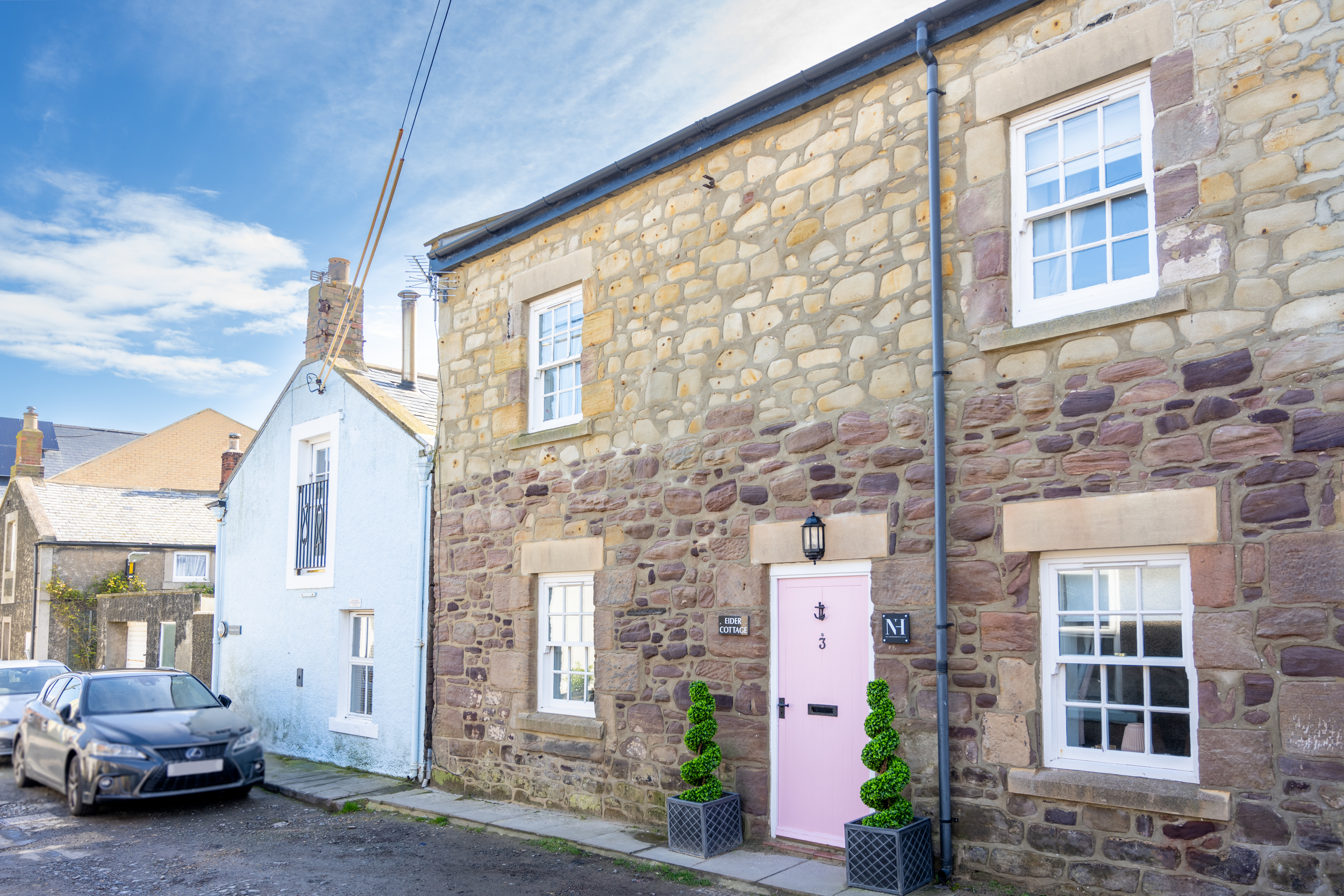 A row of stone houses with white-framed windows and a pink front door, decorated with two potted topiary trees, under a blue sky with clouds.