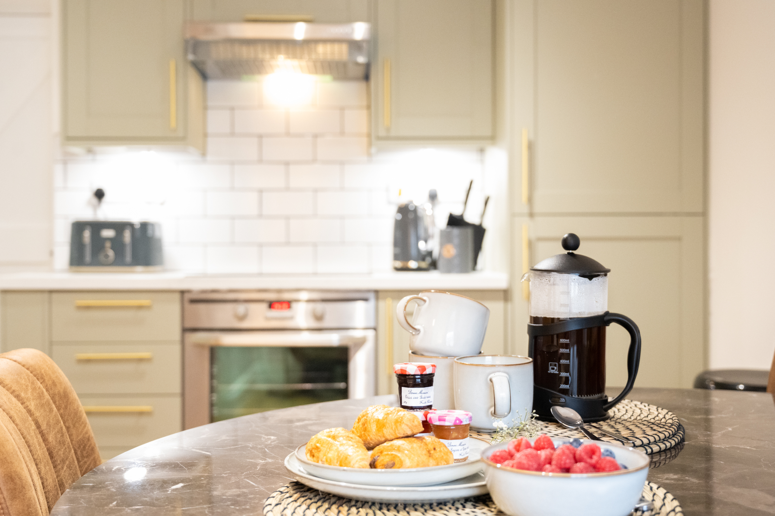 A kitchen island with croissants, jars of jam, and a cup on a plate. In the background, there is a coffee maker, bowls with raspberries and blueberries, and a white mug. The kitchen has beige cabinets, a white subway tile backsplash, and a stainless 