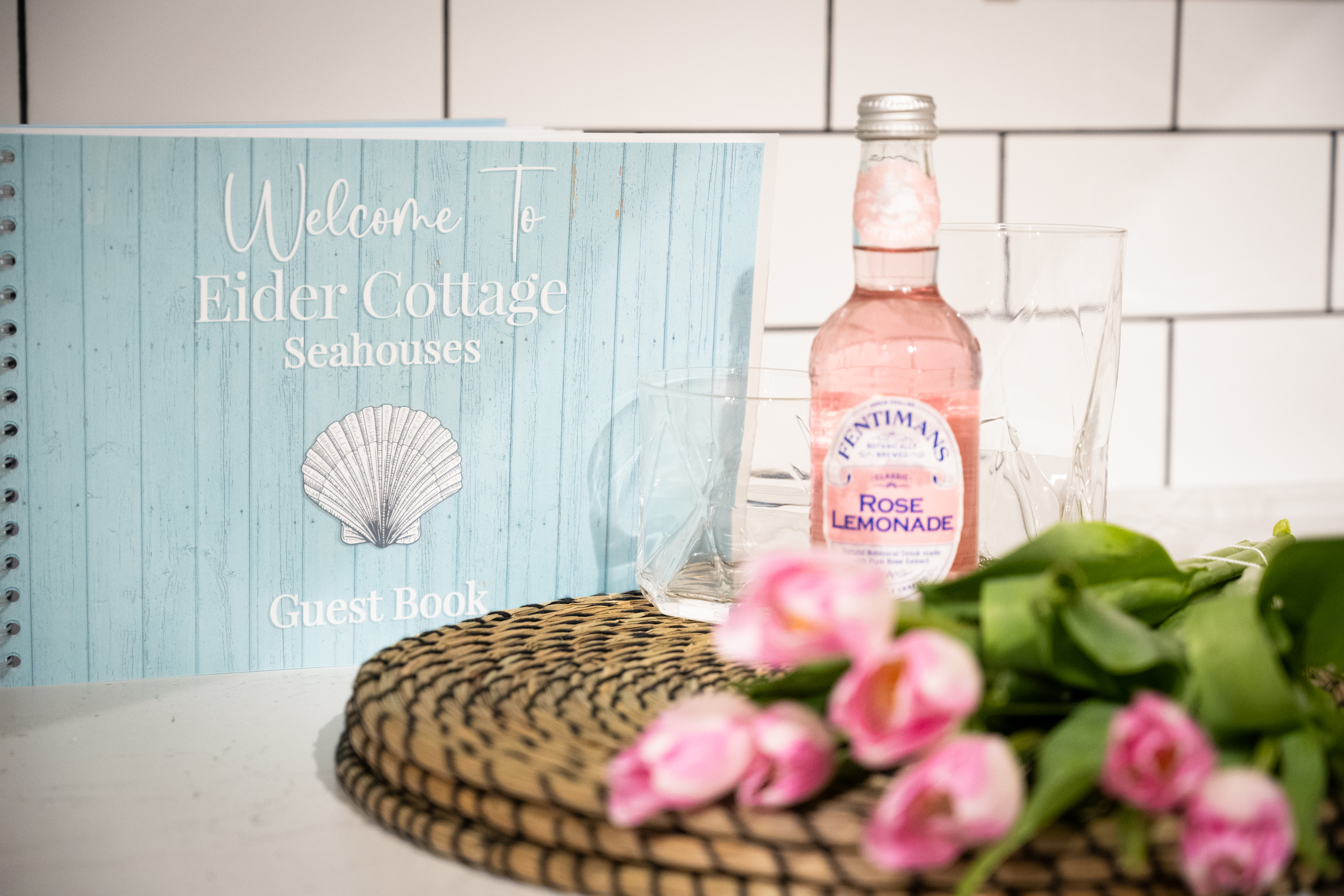 A guest book titled 'Welcome to Elder Cottage Seahouses' resting on a surface with a woven placemat in front of it. To the right, there is a pink rose lemonade bottle and empty glasses. Pink flowers and greenery are in the foreground.
