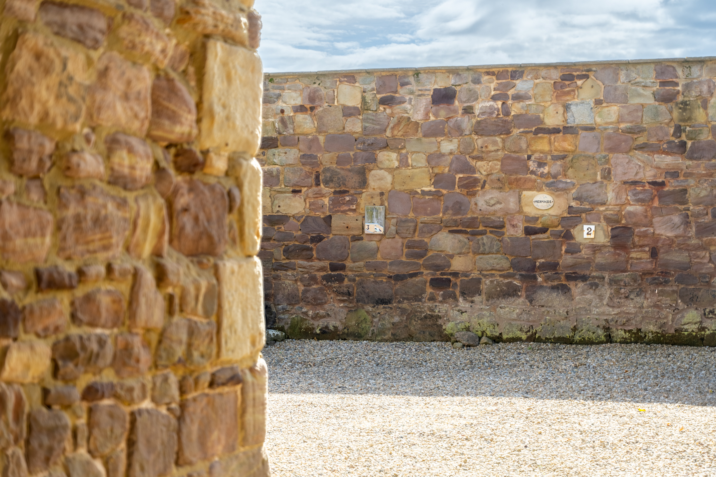 Close-up view of a brick wall, with a background stone wall under a partly cloudy sky.