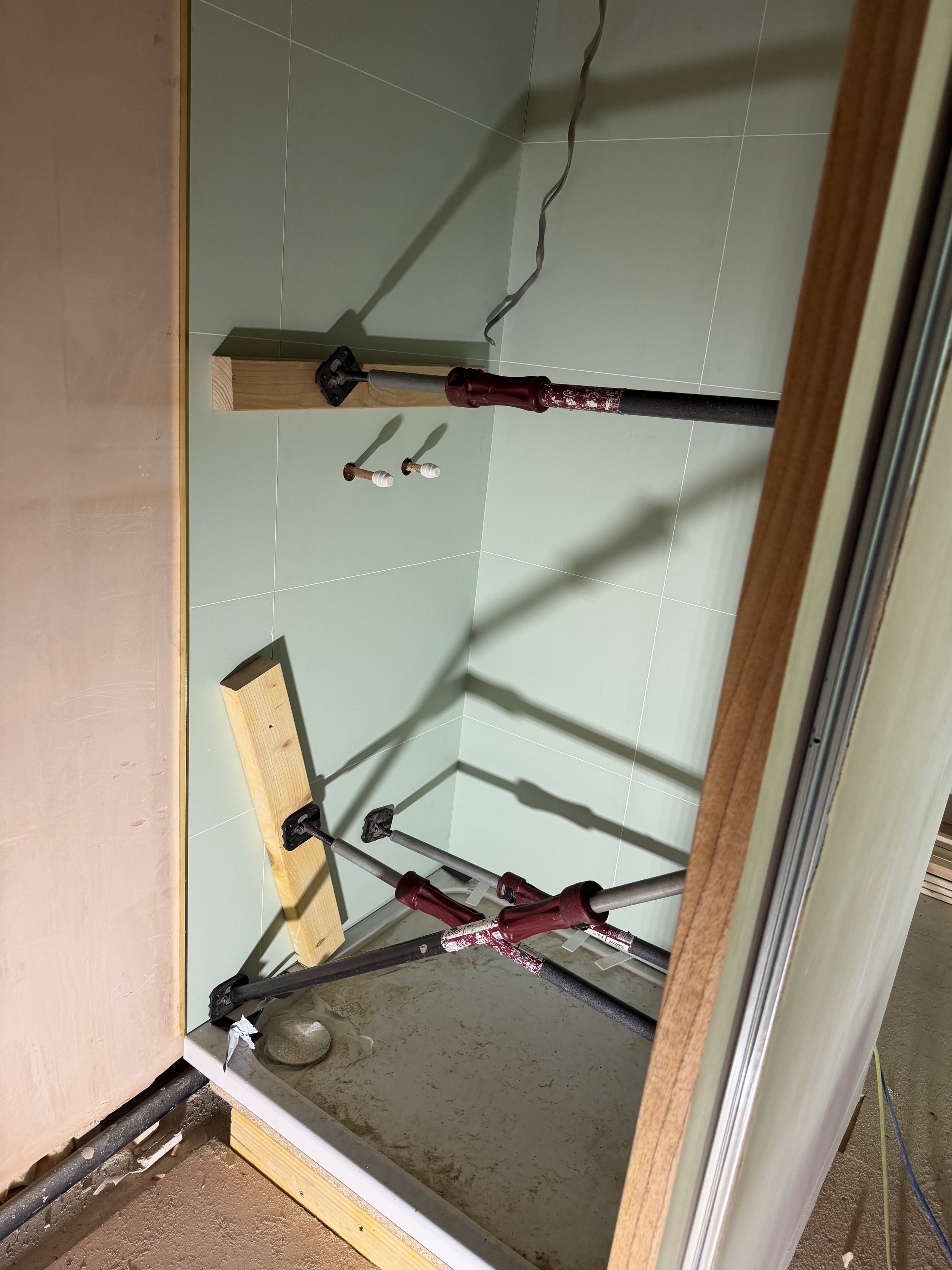 Bathroom mirror reflects a plumbing repair area with green tiled wall, pipes, tools, and wooden support blocks.