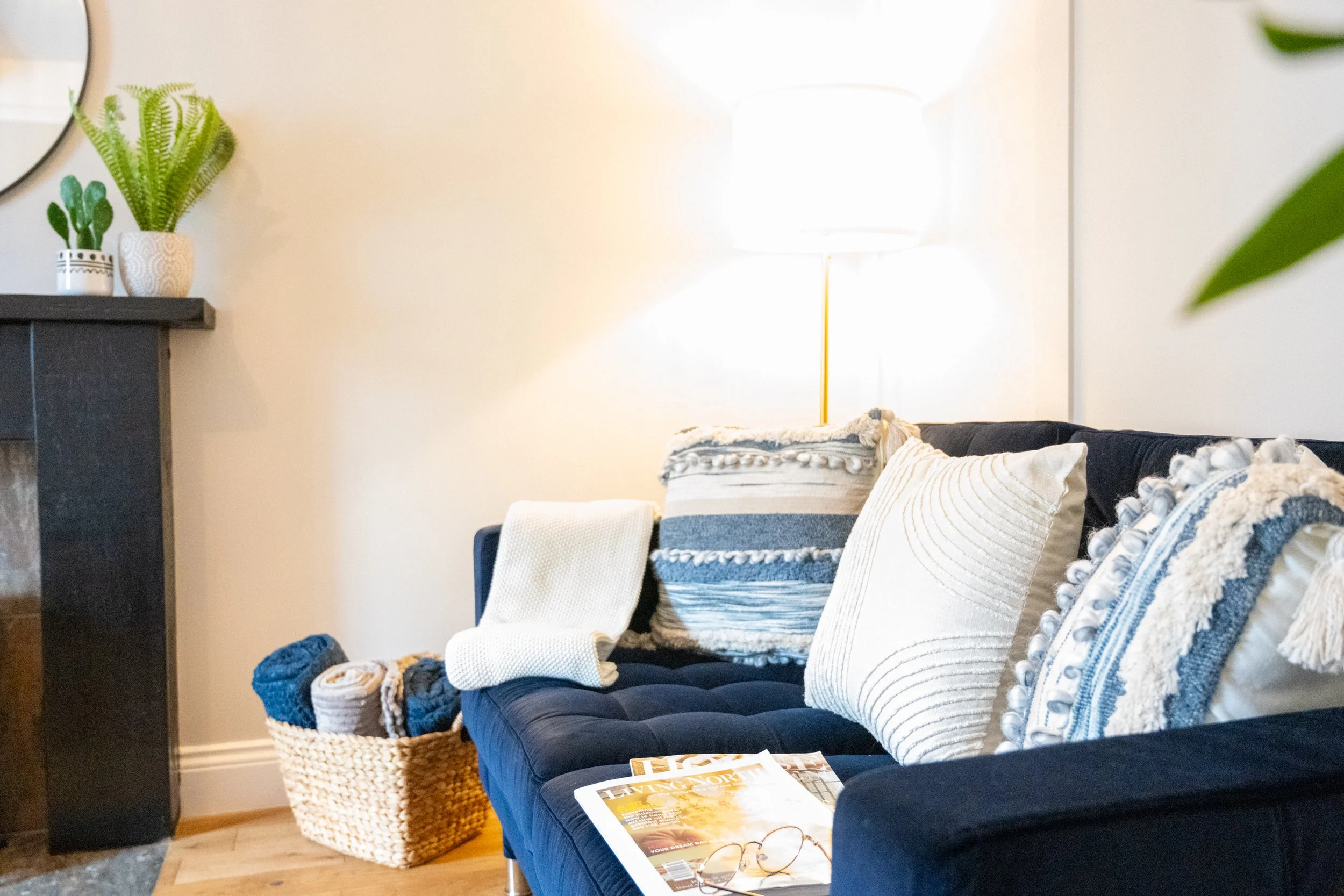 Living room with a dark blue tufted sofa decorated with pillows, a magazine and glasses on it. Wicker basket with blanket and a gray armchair. Wall with a lamp, mirror, and potted plants.