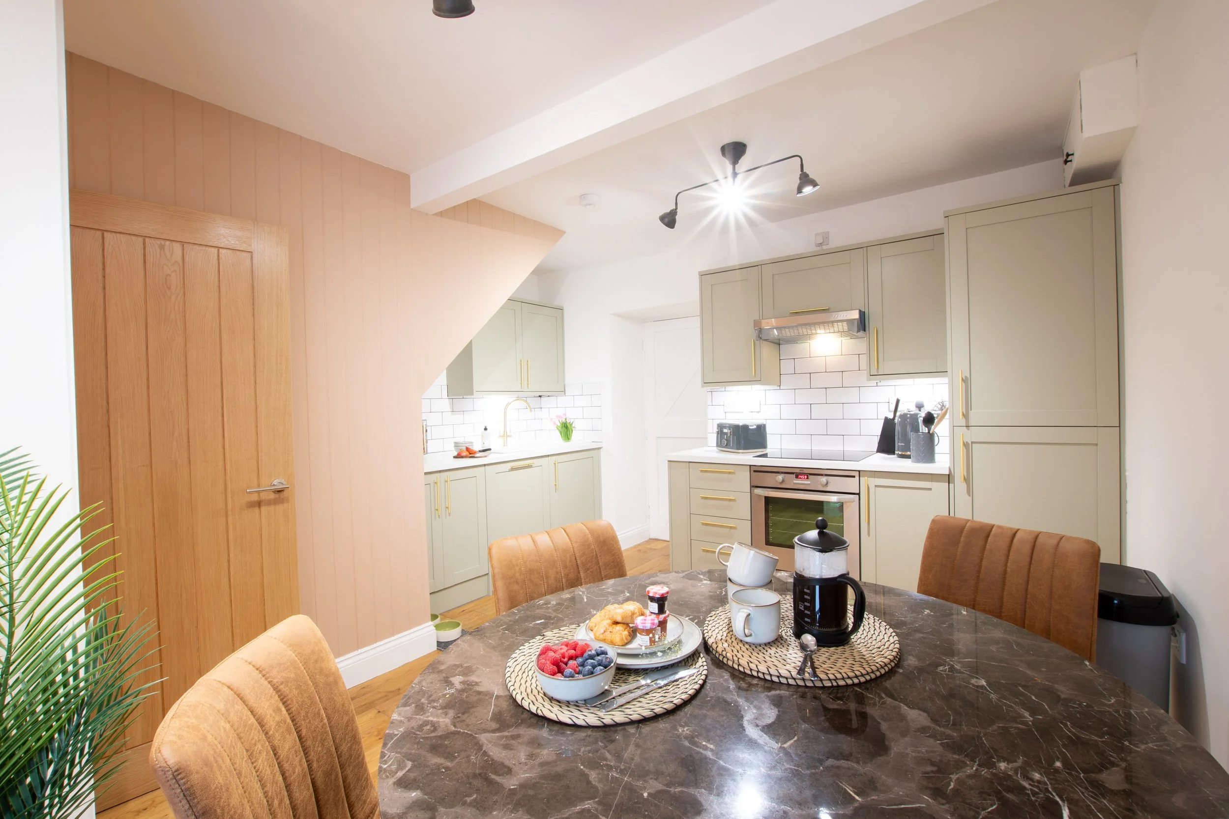 A cozy kitchen and dining area with a round marble table set with breakfast foods, surrounded by brown upholstered chairs. The kitchen features pastel green cabinets, a white tile backsplash, and window with a vase of tulips. The ceiling has black tr