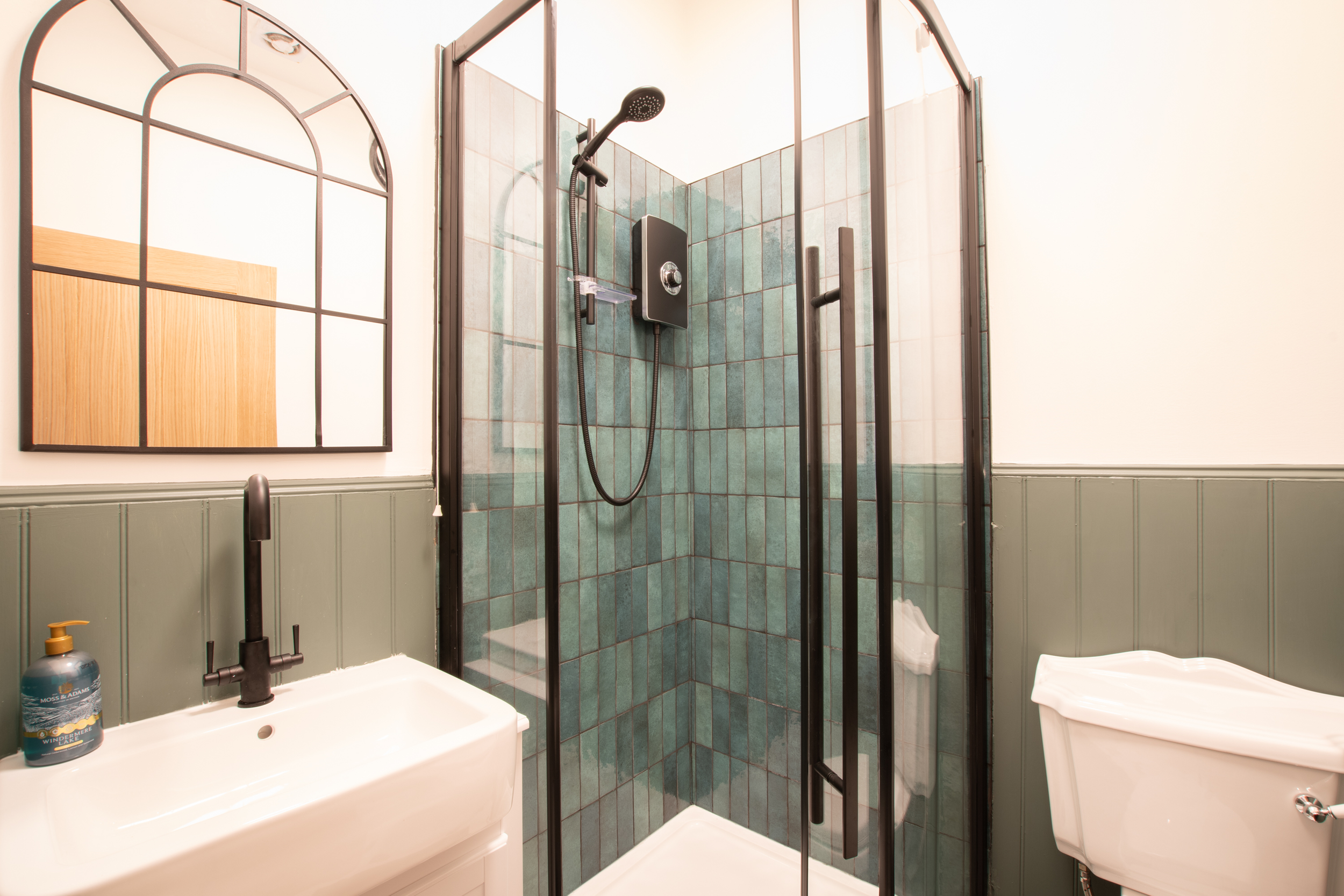 A bathroom with a glass shower enclosure, green tiles inside, a white sink with a black faucet, a mirror, a bottle of hand soap, and a white toilet.