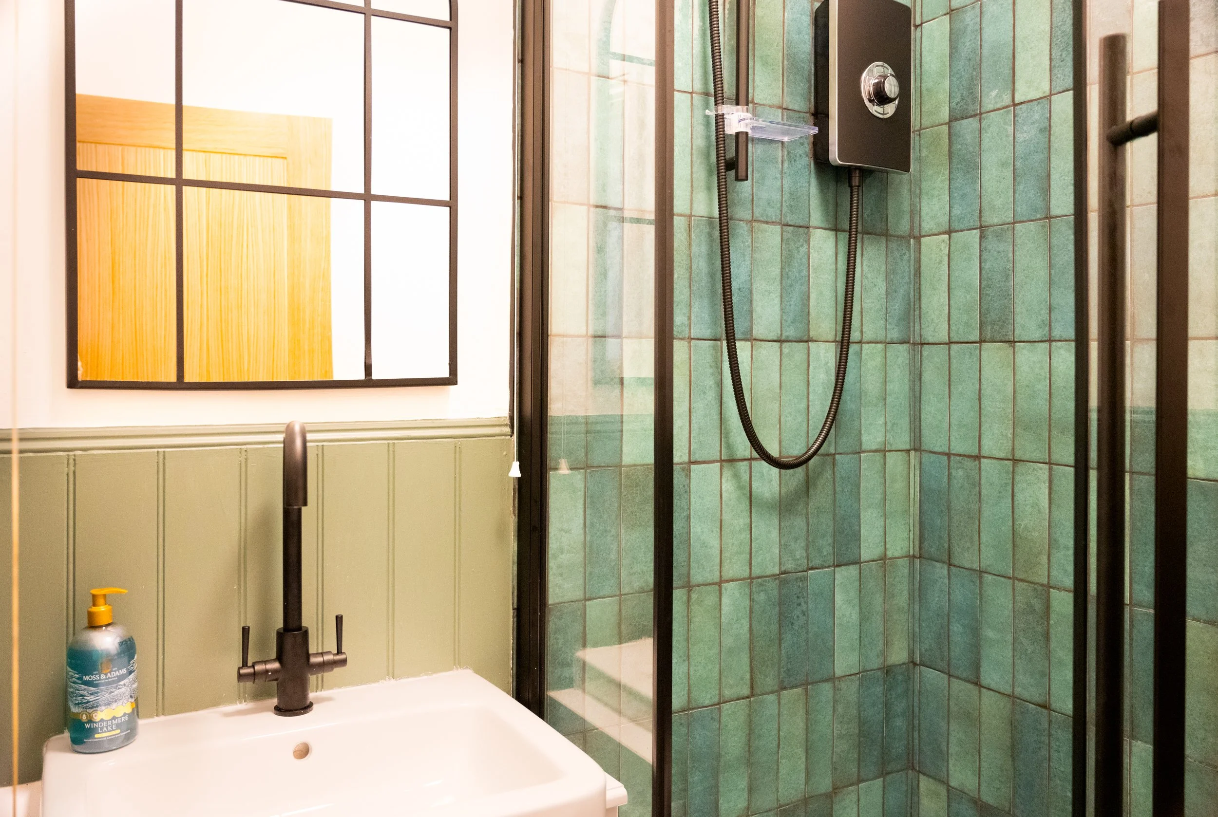 Bathroom with green tiled shower stall, black shower panel, and a white sink with a black faucet. A mirror hangs on the wall, and a bottle of shower cleaner is on the sink.