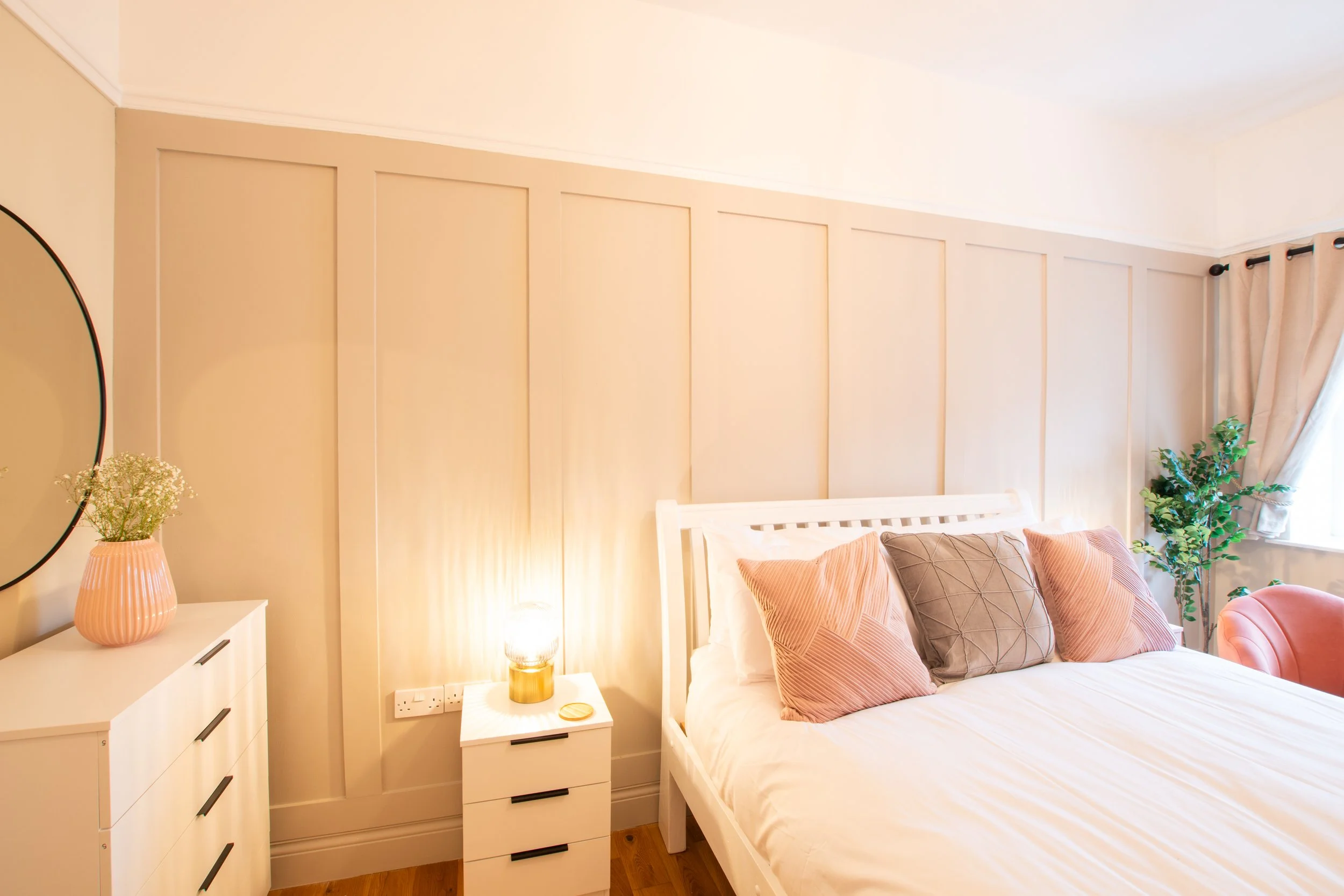 A bedroom featuring a bed with pink and gray pillows, a white bedside table with a gold lamp, a white dresser with black handles, and a window with beige curtains, a plant, and a round mirror on the wall.