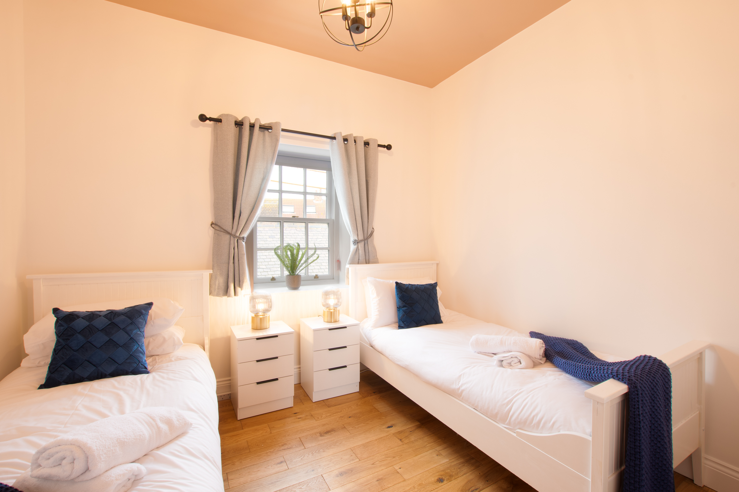 A cozy bedroom with two beds, each decorated with navy blue pillows, a window with beige curtains, a potted plant on the windowsill, and matching bedside tables with small lamps. The walls are painted light peach, and a chandelier hangs from the ceil
