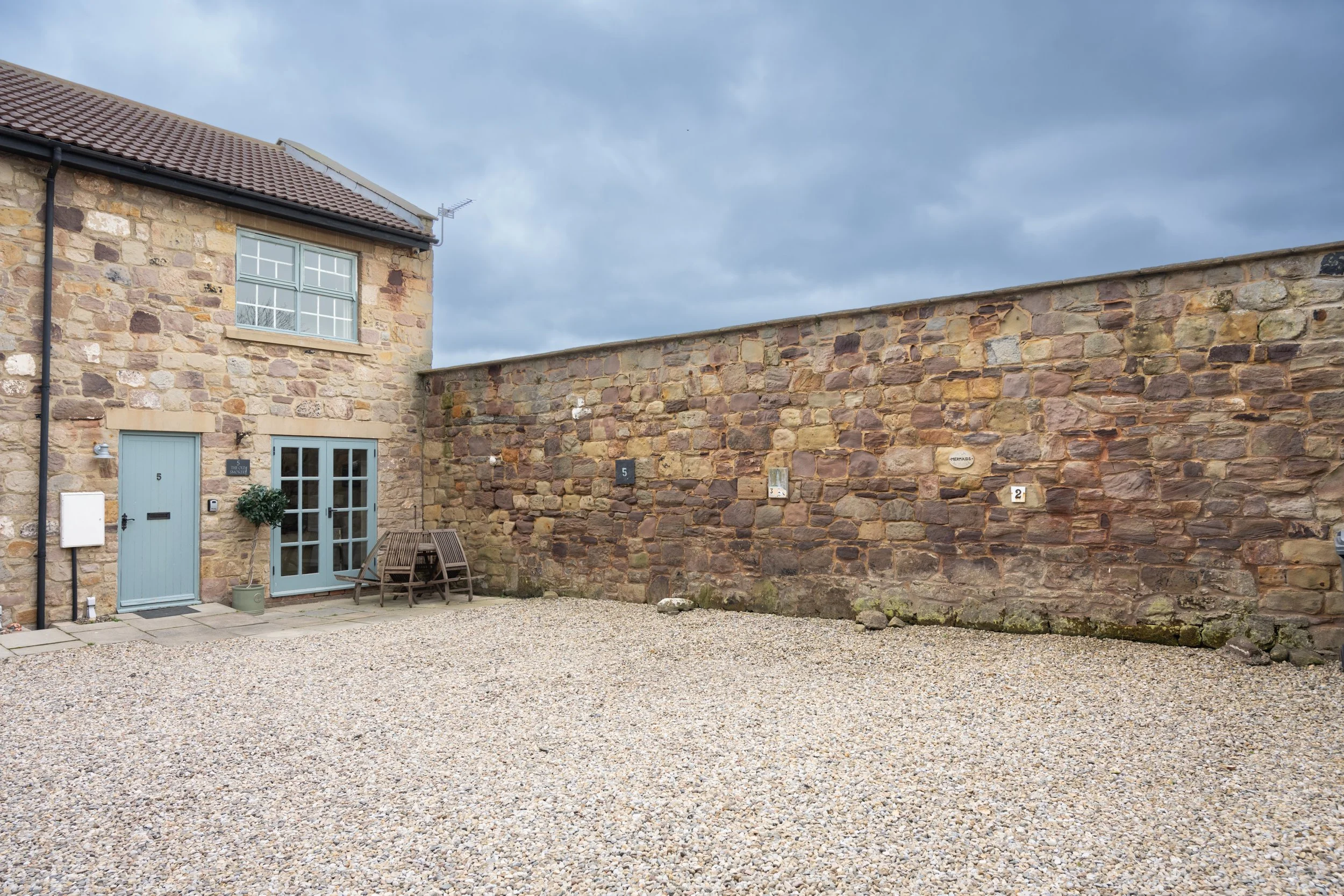A stone house with a light blue door and window frames, a small potted tree outside, and a gravel courtyard. A tall stone wall runs along the right side of the image under a cloudy sky.