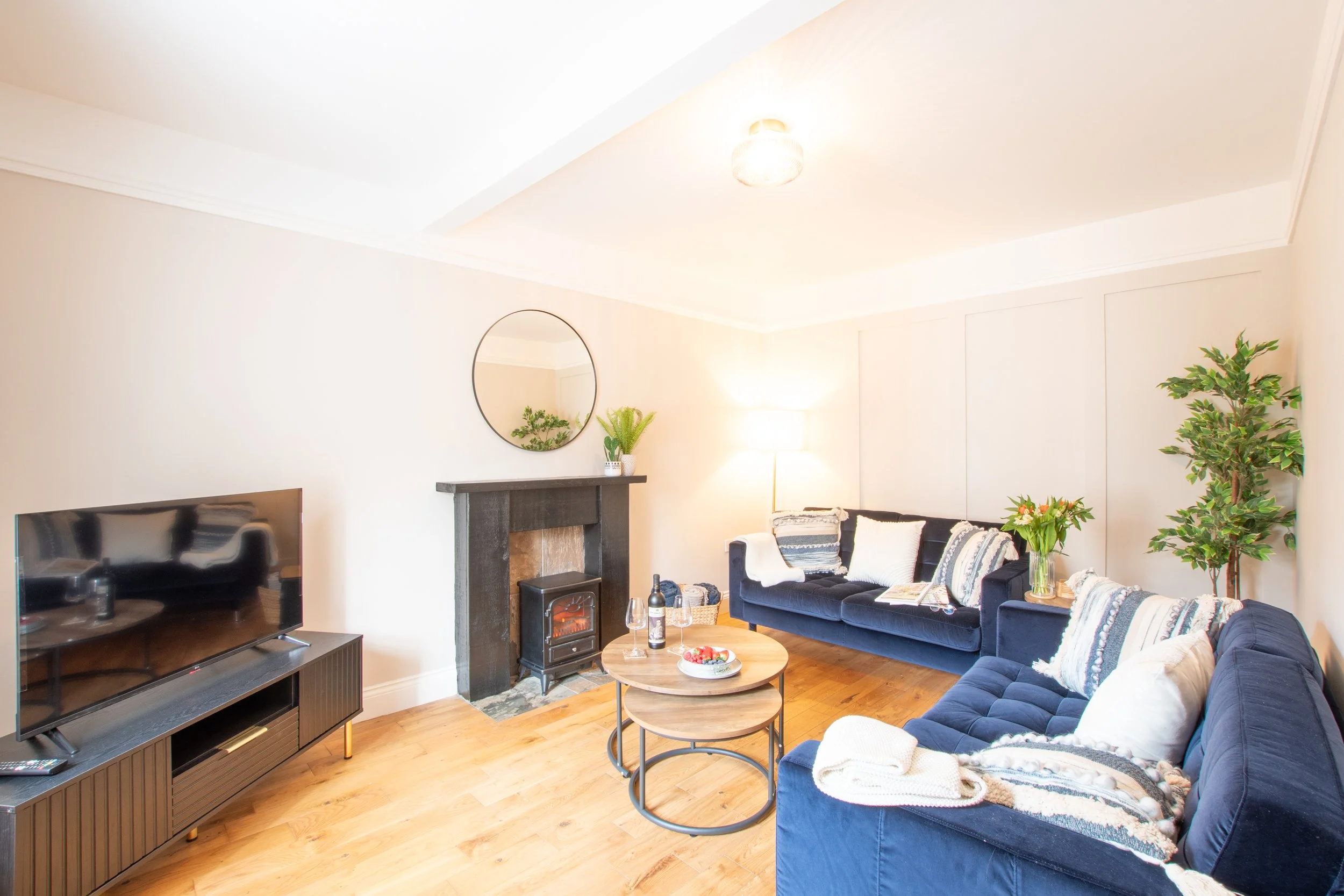 Living room with blue sofas, a television, a fireplace, a round coffee table with drinks and a bowl of strawberries, a tall plant, a mirror, and a lamp.